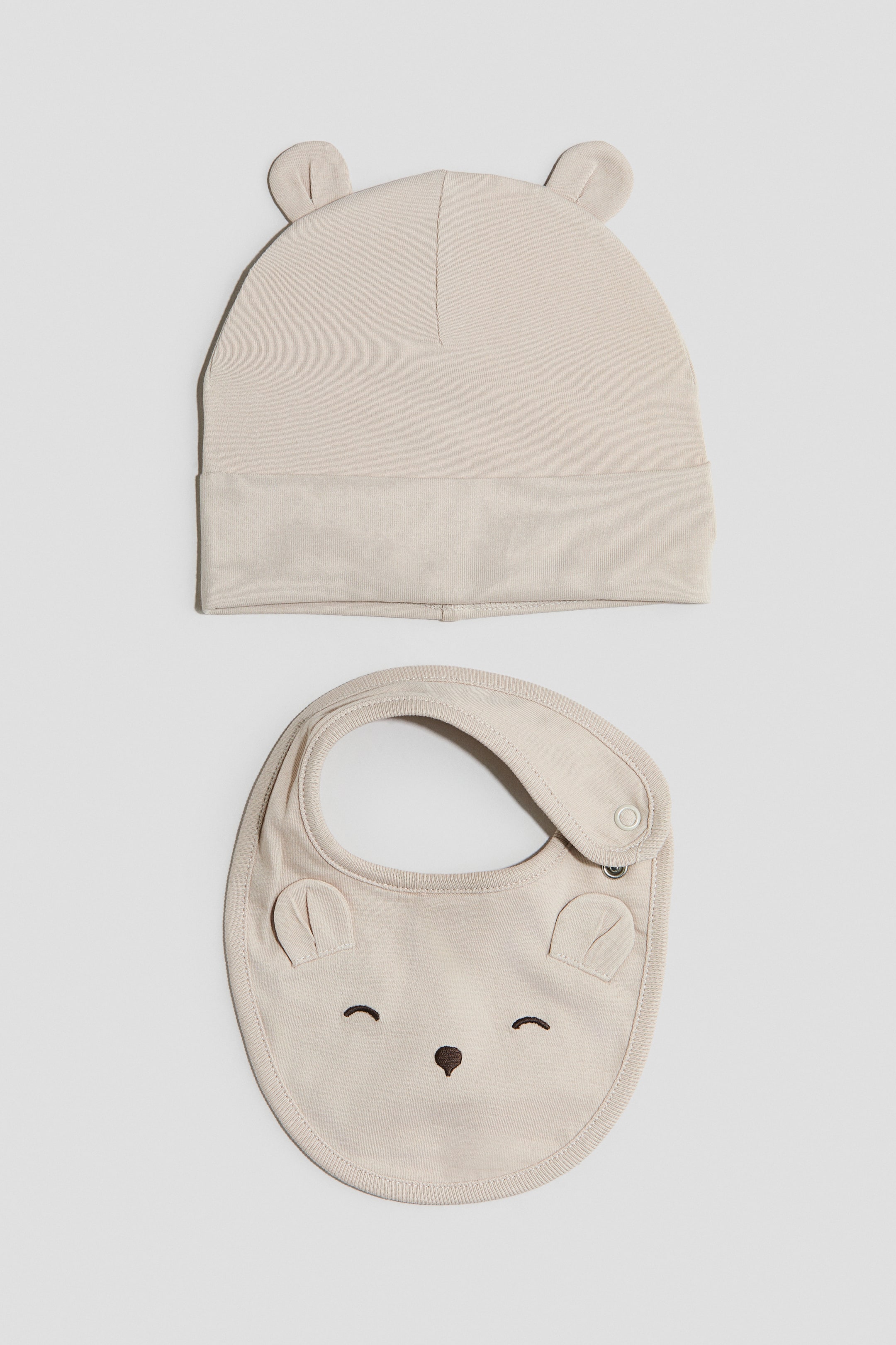 Ver imagem maior: Um gorro de bebé bege claro e um babete a condizer estão dispostos na horizontal. O gorro apresenta dois pequenos detalhes arredondados em forma de orelhas no topo, enquanto o babete exibe dois pequenos detalhes de tecido arredondados em forma de orelhas, olhos fechados bordados e um pequeno nariz, além de um fecho de pressão na tira do pescoço.