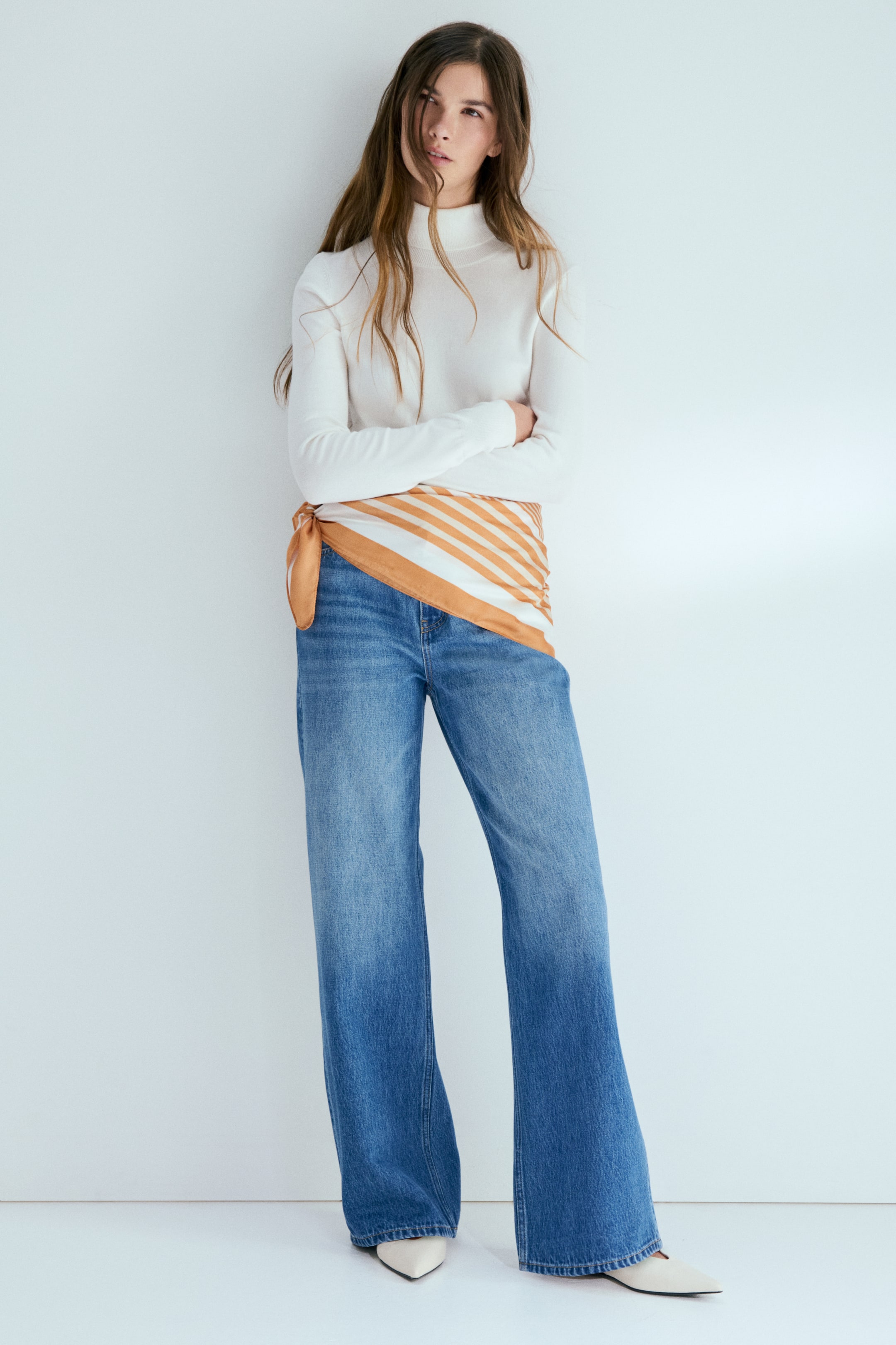 View larger image: A white long-sleeve turtleneck top is paired with blue wide-leg jeans that have a faded wash. An orange and white diagonally striped scarf is tied around the waist, and cream pointed-toe flats complete the look.