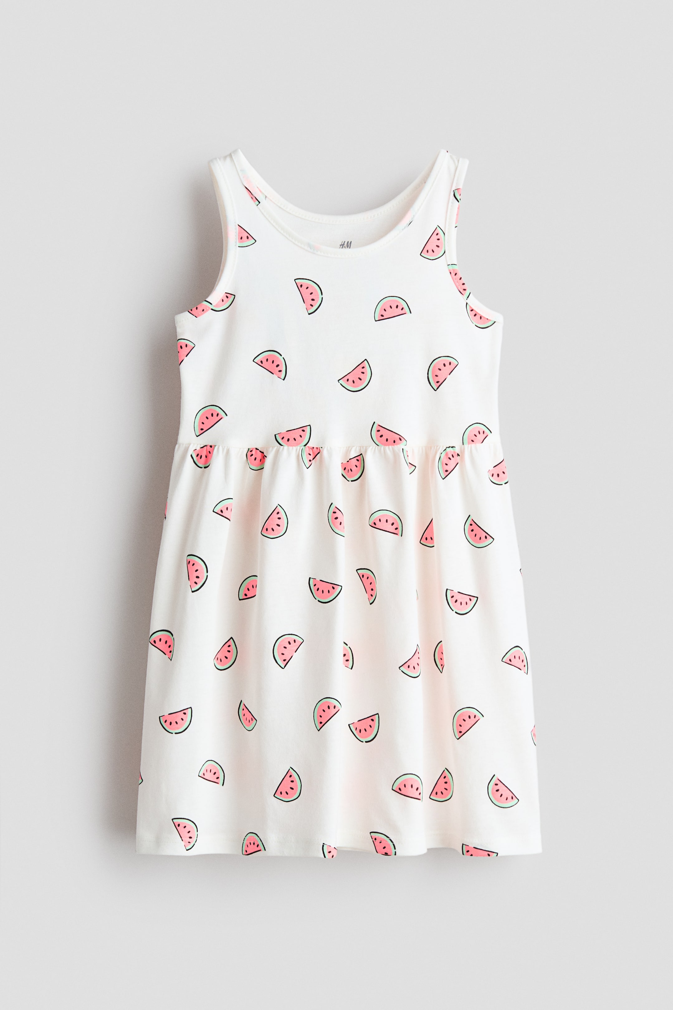 Ingrandisci l'immagine: Un abito senza maniche è mostrato frontalmente, realizzato in tessuto jersey bianco con un motivo a fette di anguria su tutta la superficie. È progettato con uno scollo rotondo, spalline in stile canotta e presenta un'arricciatura alla cucitura della vita, che conduce a una gonna leggermente svasata.