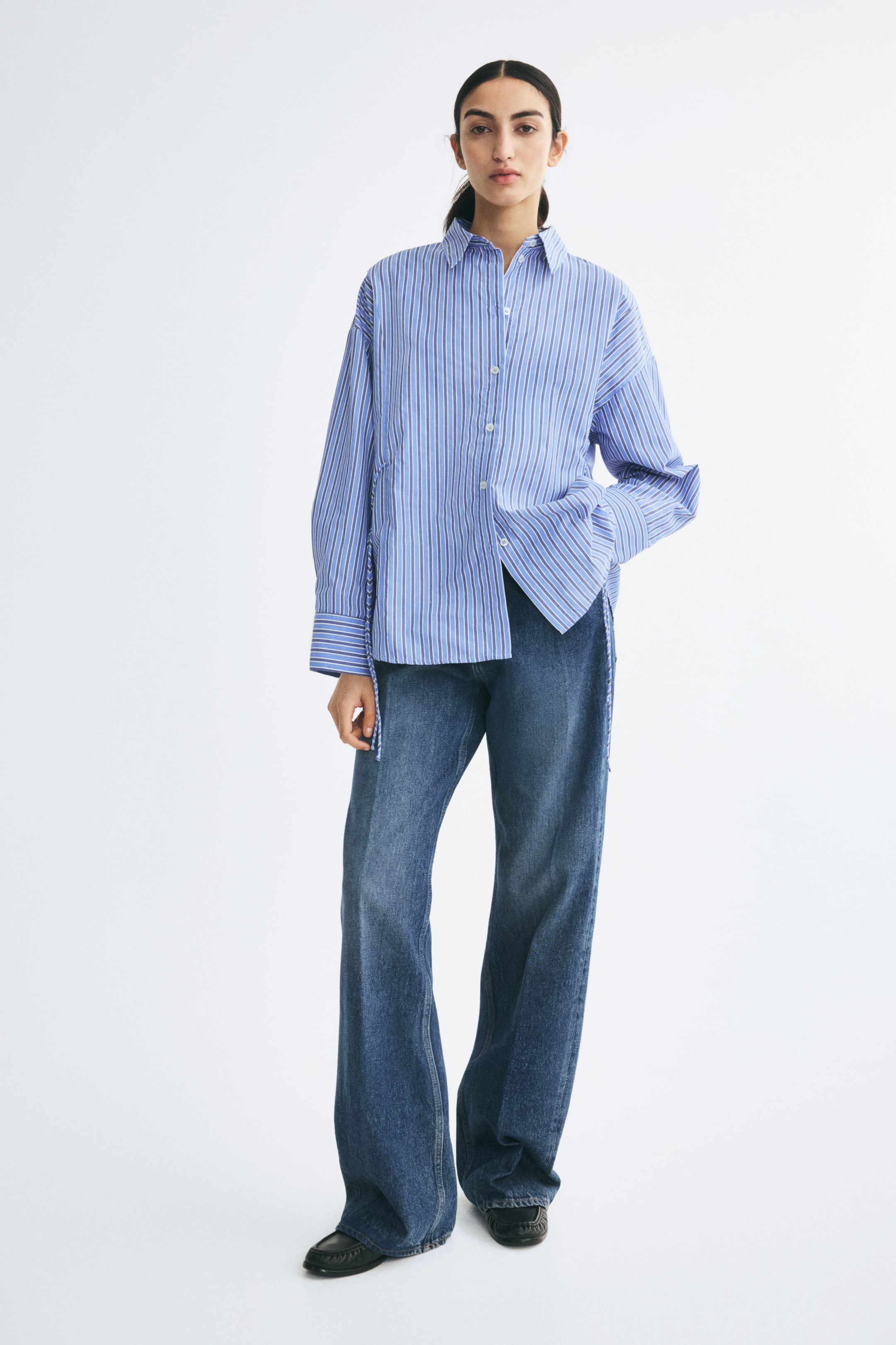 View larger image: A blue and white striped woven shirt featuring a collar, front button closure, dropped shoulders, and narrow ties at the waist is worn by the model. The shirt has long sleeves with wide button cuffs and is styled loose over wide-leg blue denim jeans.