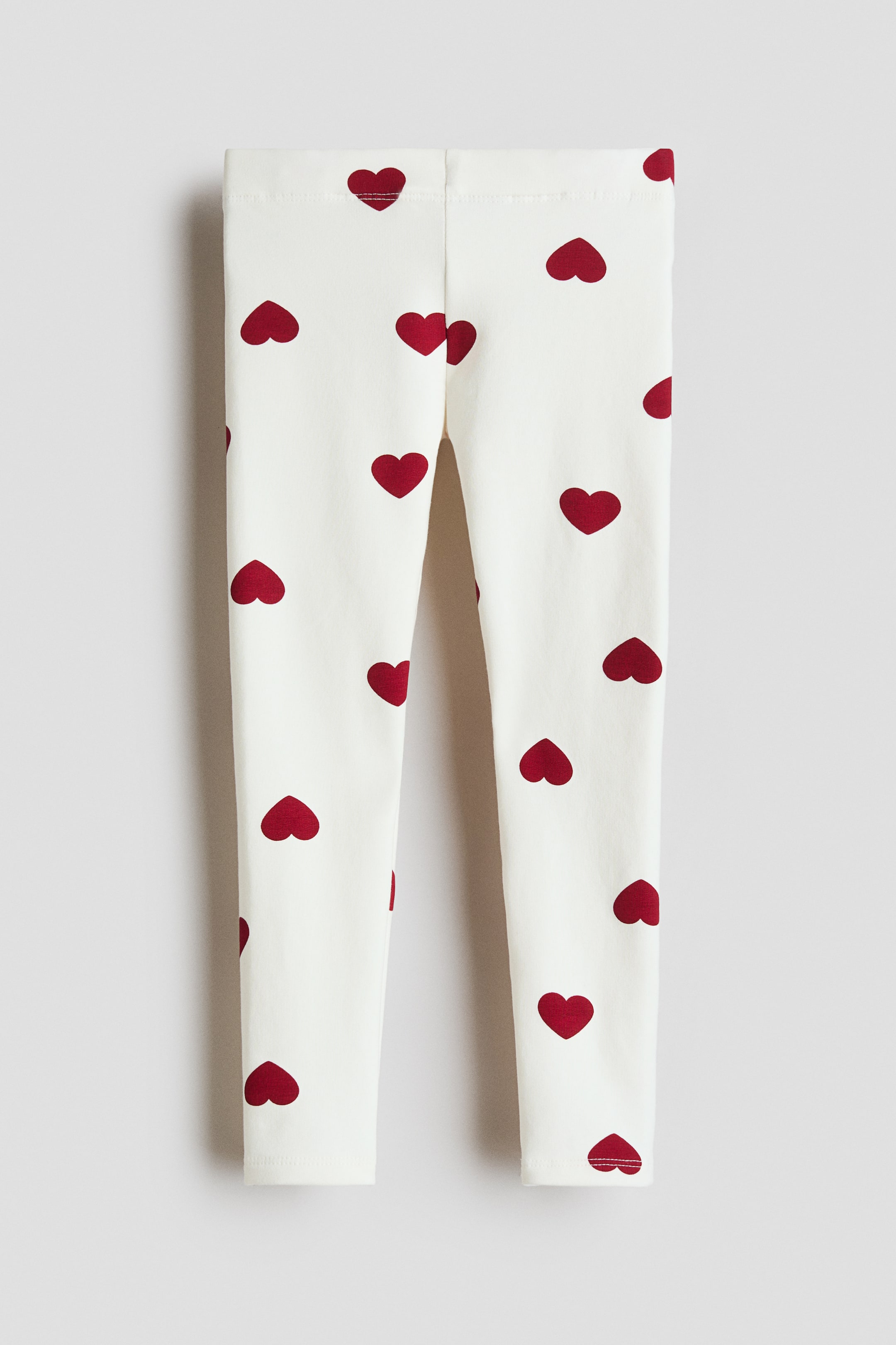 View larger image: Leggings are presenting from the front, featuring an off-white color with a scattered red heart pattern. They are showing a flat waistband and full-length legs.