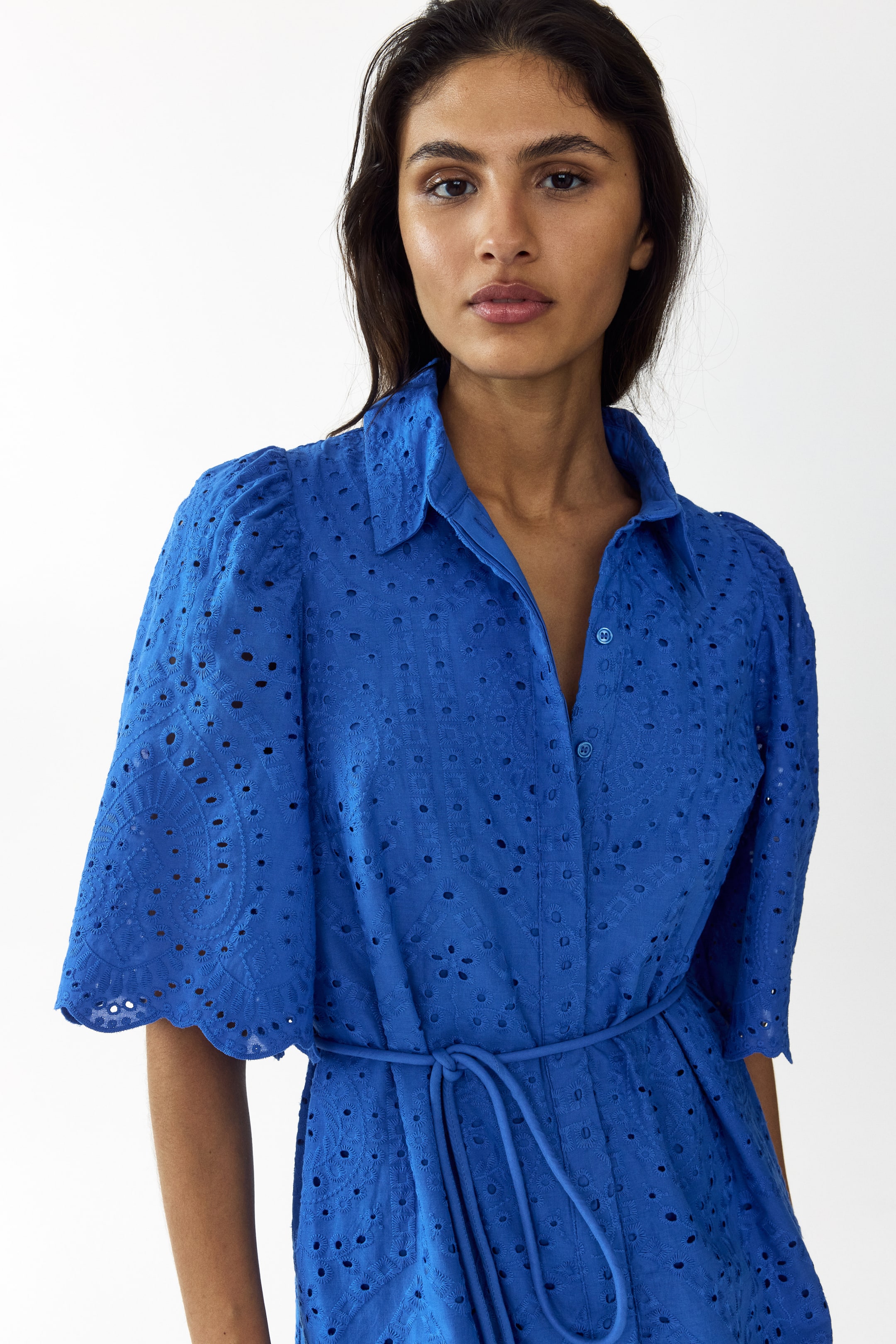 View larger image: A blue eyelet lace shirt dress features a pointed collar, a button-down front, and puffed short sleeves with scalloped edges. A matching tie-belt cinches the waist of the dress.