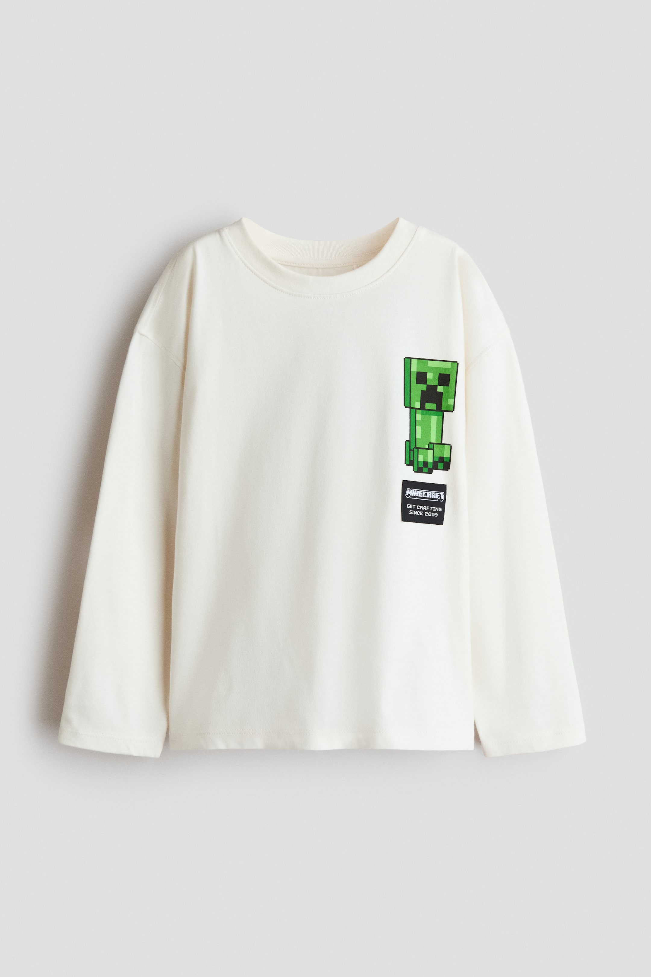 View larger image: A cream long-sleeved crewneck top features a pixelated green Minecraft Creeper character print on the left chest, with a black rectangular patch displaying "MINECRAFT GET CRAFTING SINCE 2009" in white text below it. The top also showcases a small side slit on the right side of its straight bottom hem.