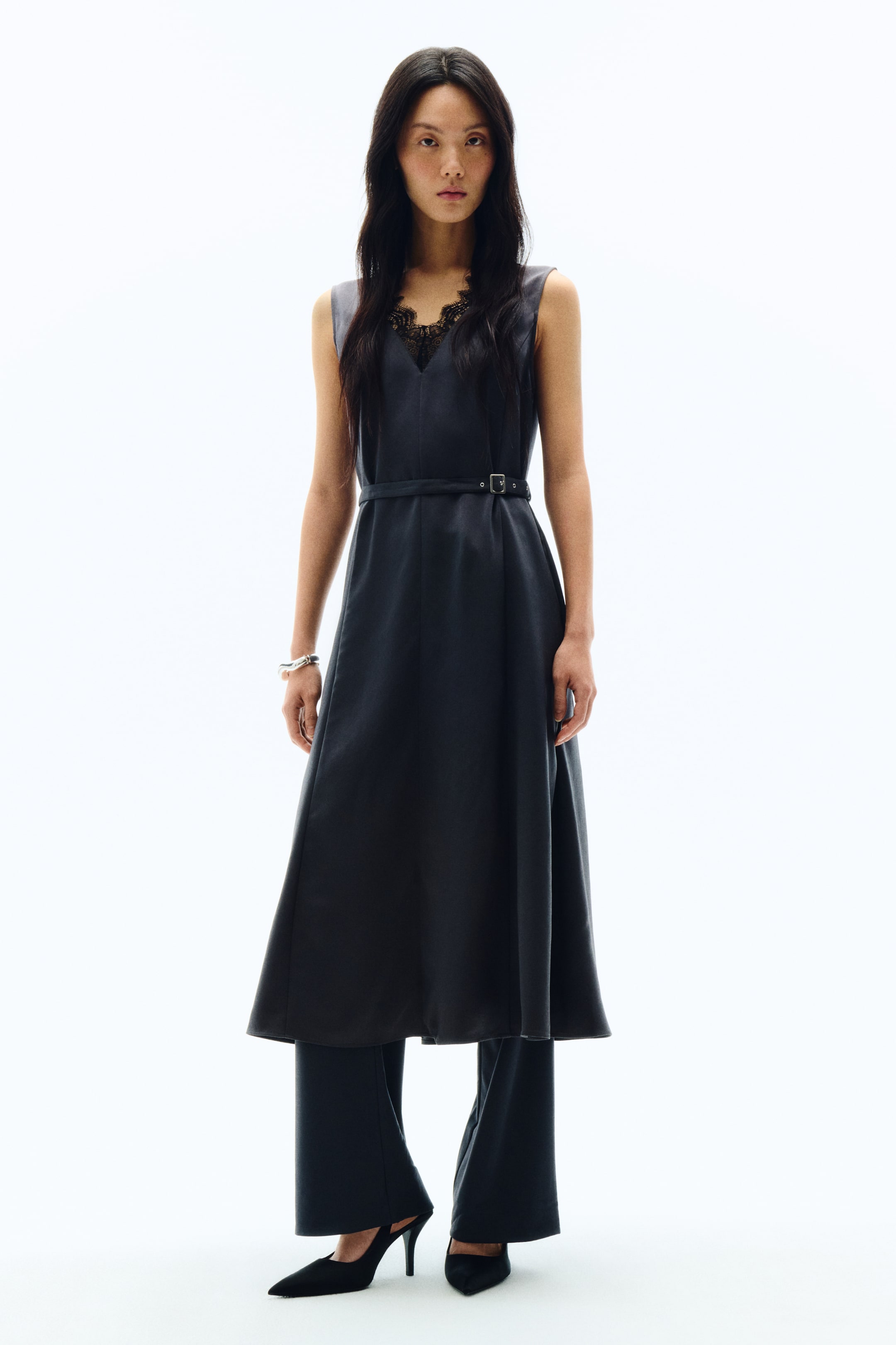 拡大画像を表示: ダークグレーのノースリーブAラインミディ丈ワンピース。Vネックラインからはブラックレースがさりげなく覗き、細身のベルトがウエストをすっきりとマークしています。ワンピースの下には同系色のワイドレッグパンツをレイヤードし、足元にはブラックのポインテッドトゥスリングバックヒールが覗いています。