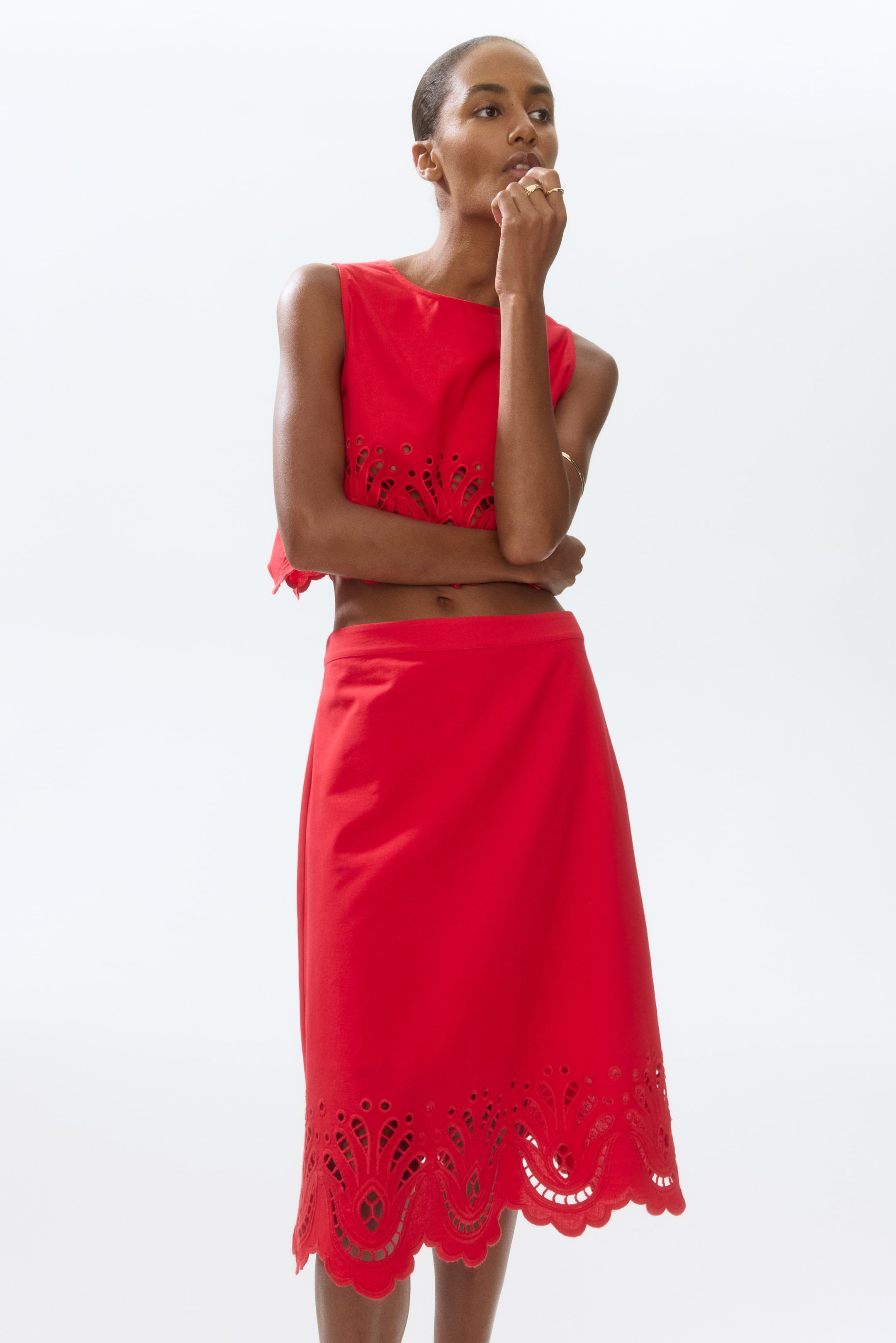 View larger image: A red sleeveless crop top showcases intricate eyelet detailing along its front bottom edge. Complementing this, the red midi skirt features a broad band of eyelet patterns and a scalloped hem at its lower edge.