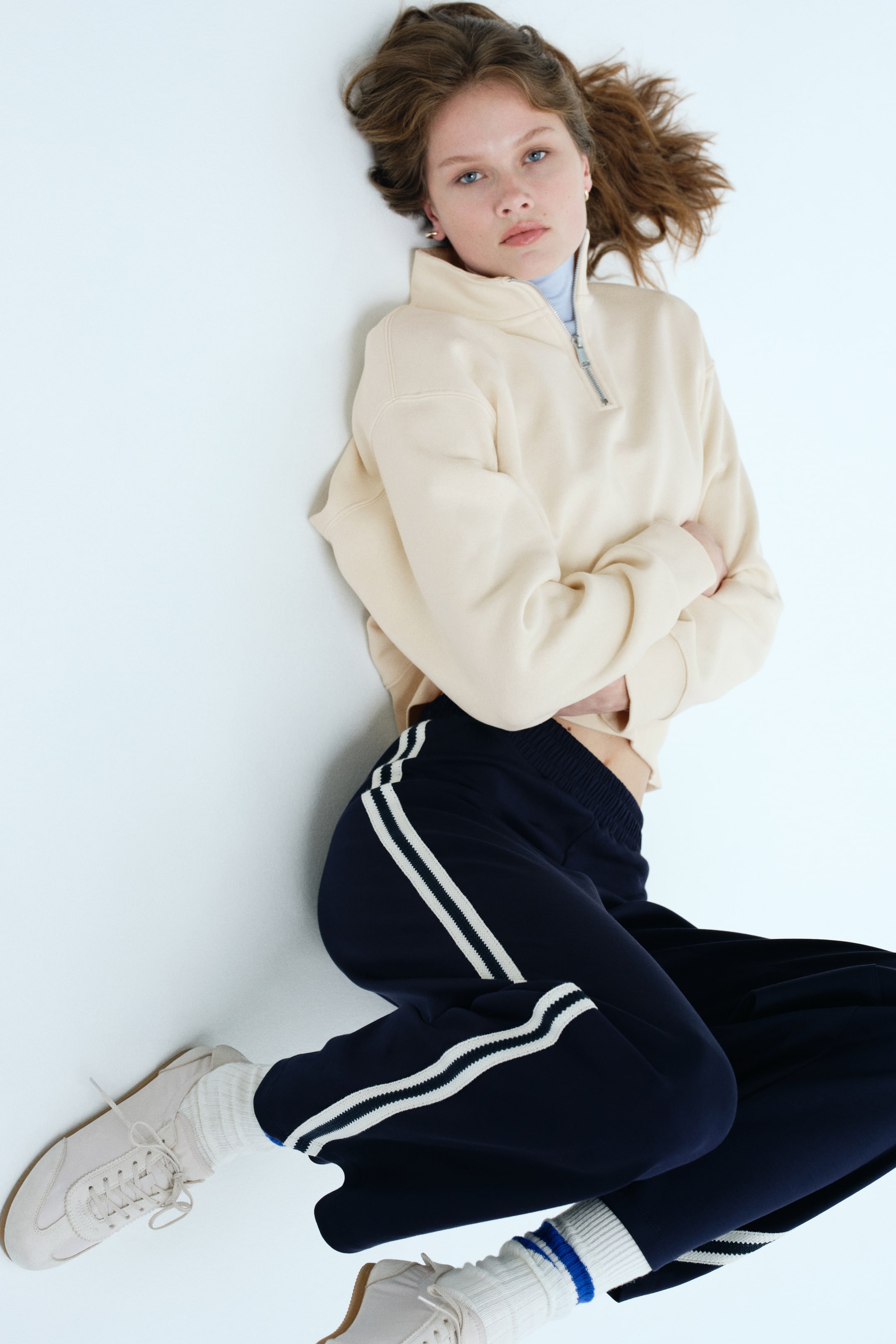 Voir une image plus grande: Un sweatshirt quarter-zip beige pâle présente un col montant avec une fermeture éclair argentée et des poignets côtelés, porté sur un col roulé bleu pâle. Un pantalon de survêtement bleu marine affiche deux bandes blanches le long des coutures extérieures et des bas côtelés avec une bande blanche, des chaussettes blanches étant visibles en dessous.
