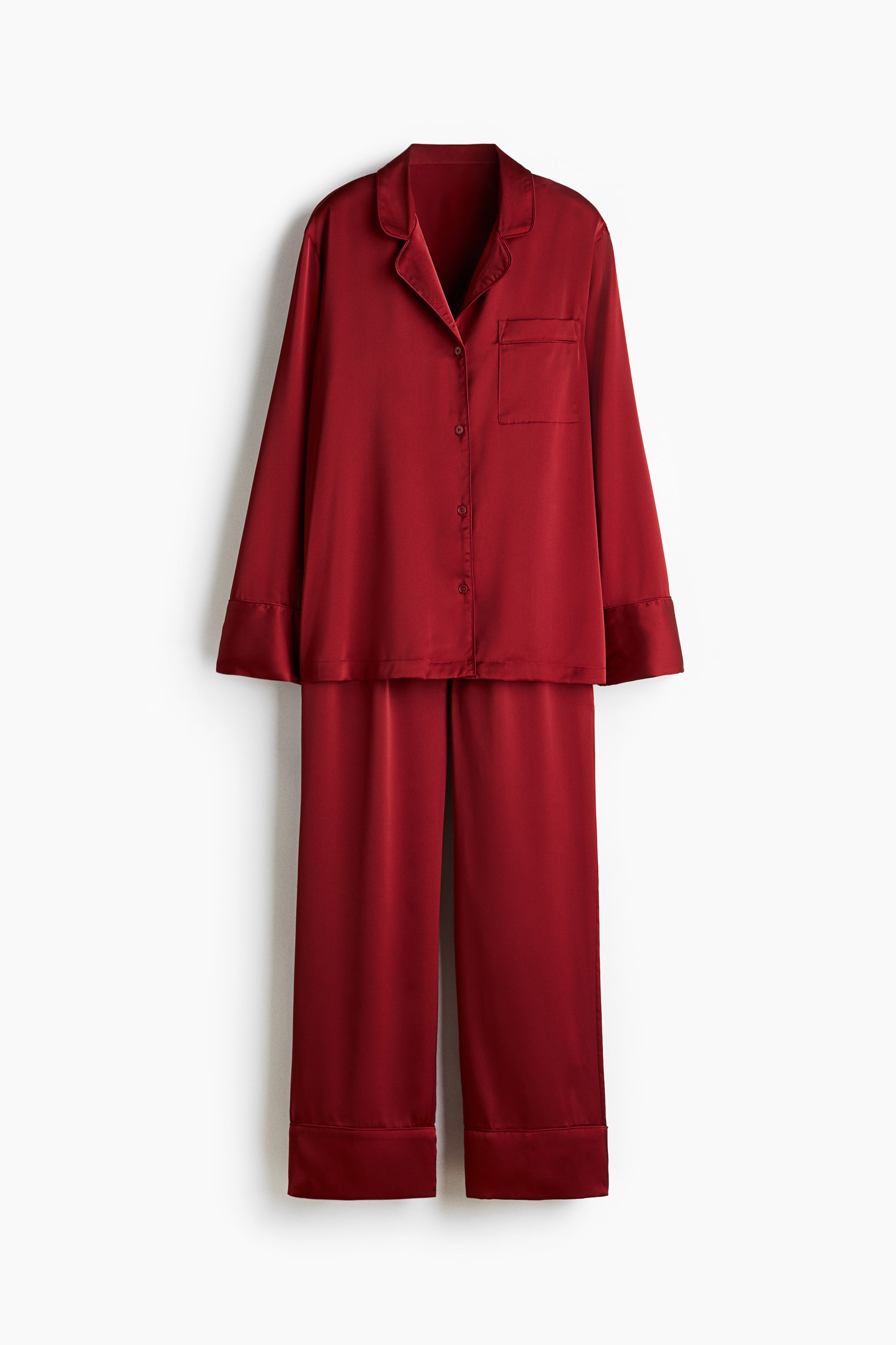 View larger image: A red pyjama set is presenting flat, front-facing, made from softly draping satin. The shirt is featuring a resort collar, buttons down the front, an open chest pocket, and long sleeves with wide cuffs. The matching bottoms are featuring wide legs with cuffed hems.