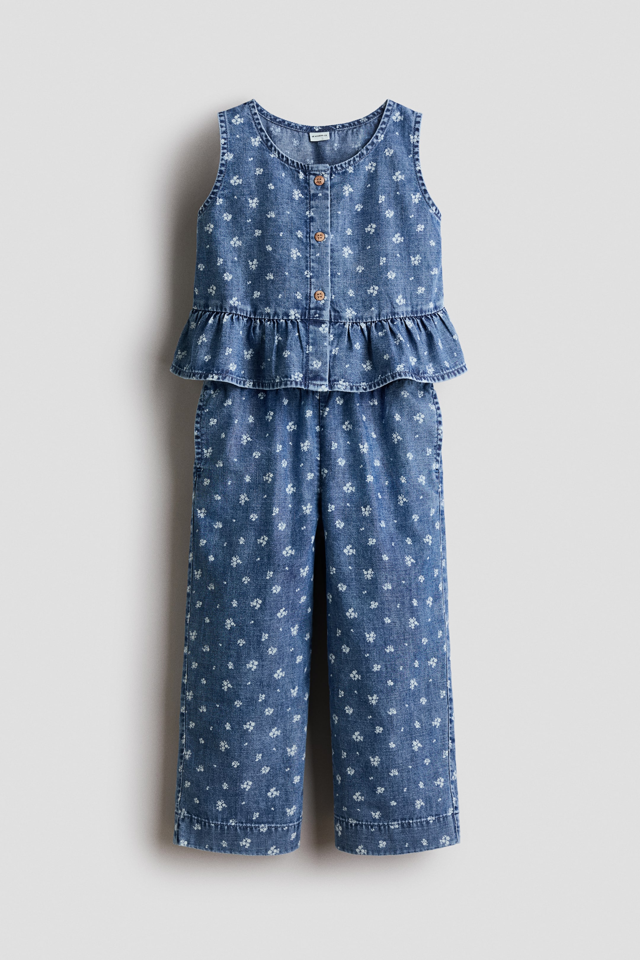 View larger image: A child's two-piece blue denim outfit is displaying a white floral print, presented facing FRONT. The sleeveless top is featuring a round neckline, a button-front placket with wooden-look buttons, and a ruffled peplum hem, while coordinating wide-leg trousers are completing the set.