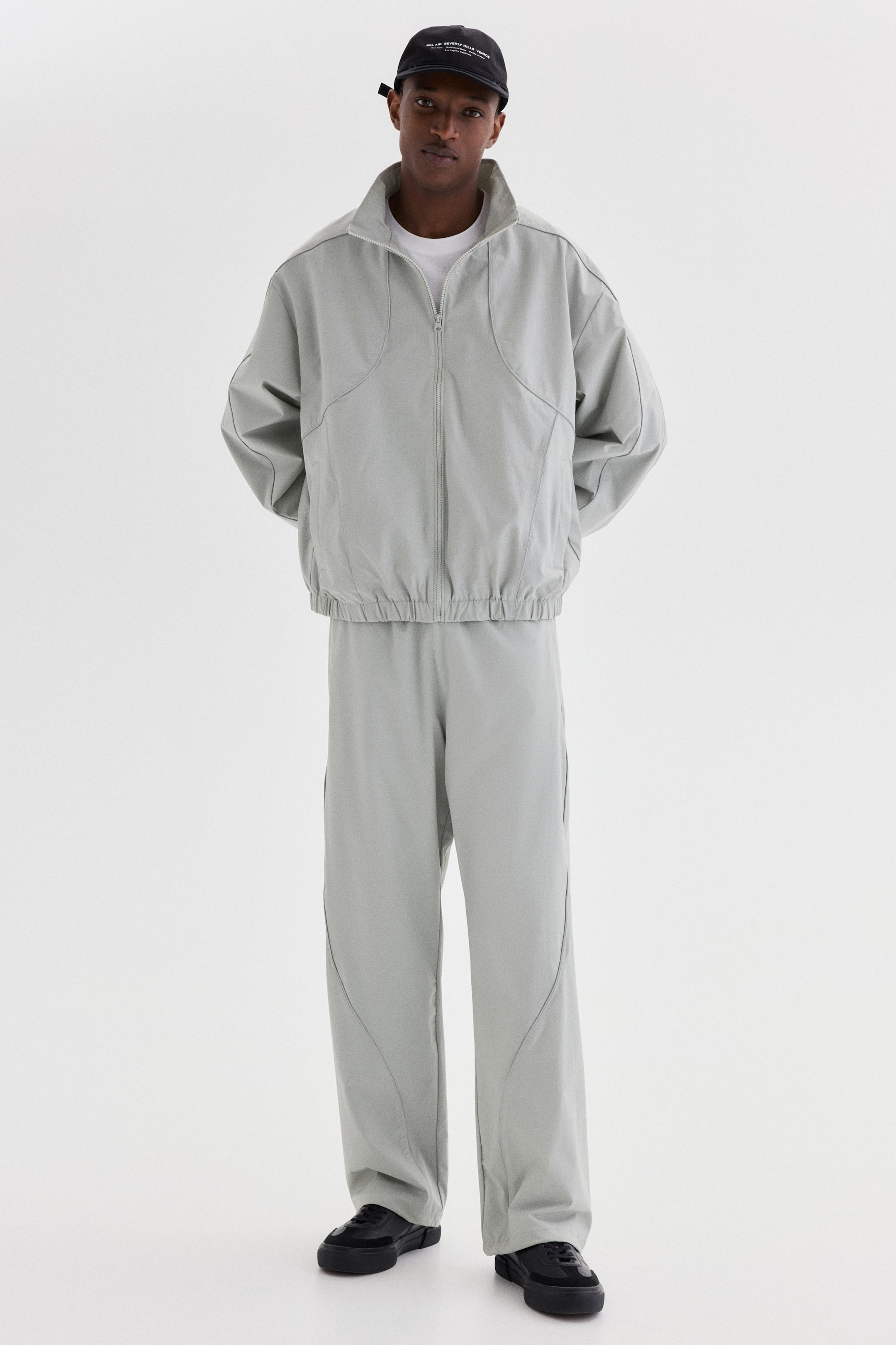 查看大圖: 淺灰色配套外套與闊腿褲，以寬鬆版型呈現獨特弧形縫線細節。外套採用立領、全開式拉鏈前襟，以及彈性袖口與下擺；配套闊腿褲在褲腿亦展現相同弧形縫線。