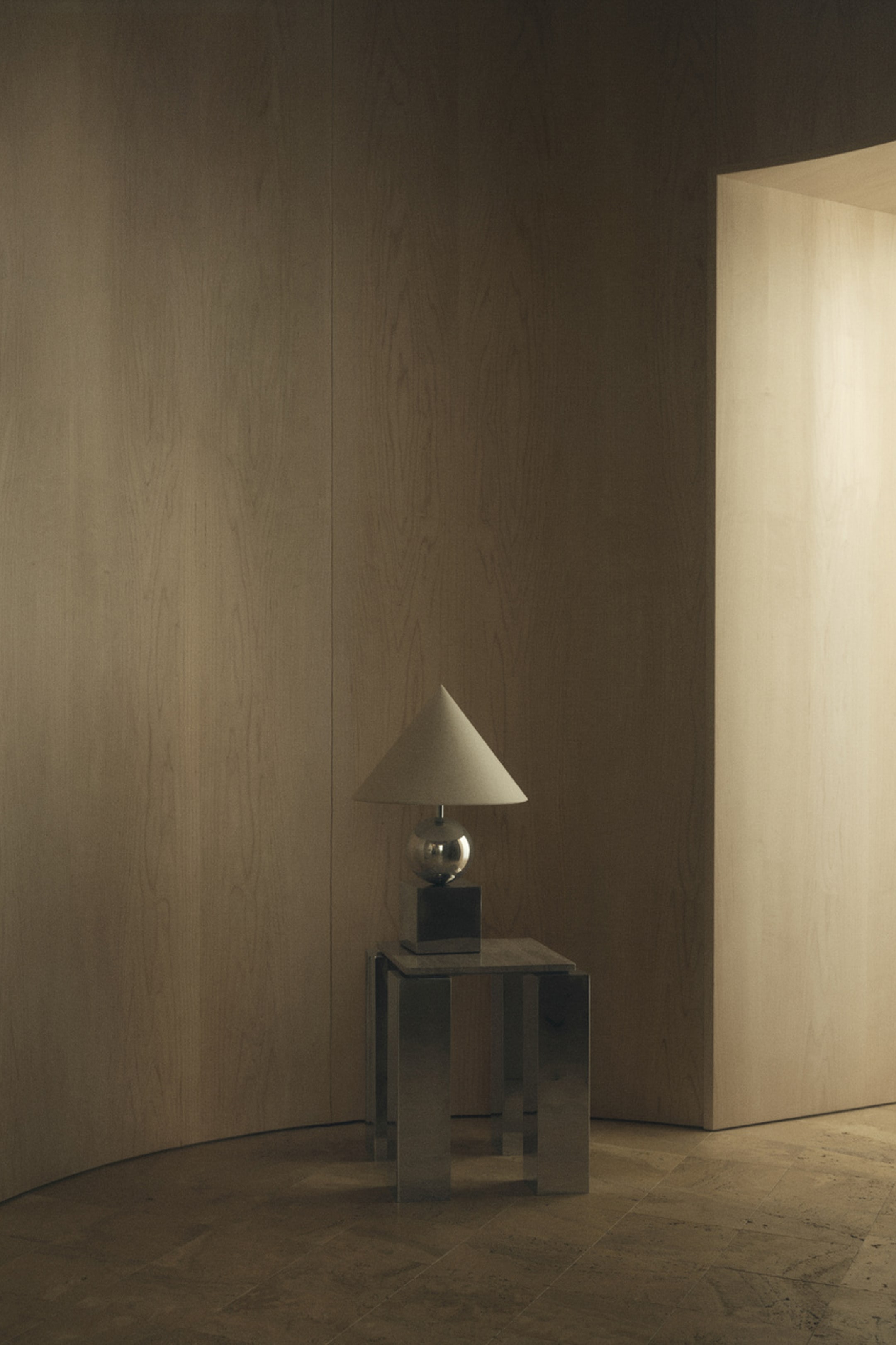 View larger image: A side table stands with its front facing forward, featuring a square tabletop in textured travertine stone supported by four chunky, reflective stainless steel block legs. A metallic table lamp with a conical shade rests on the stone surface.