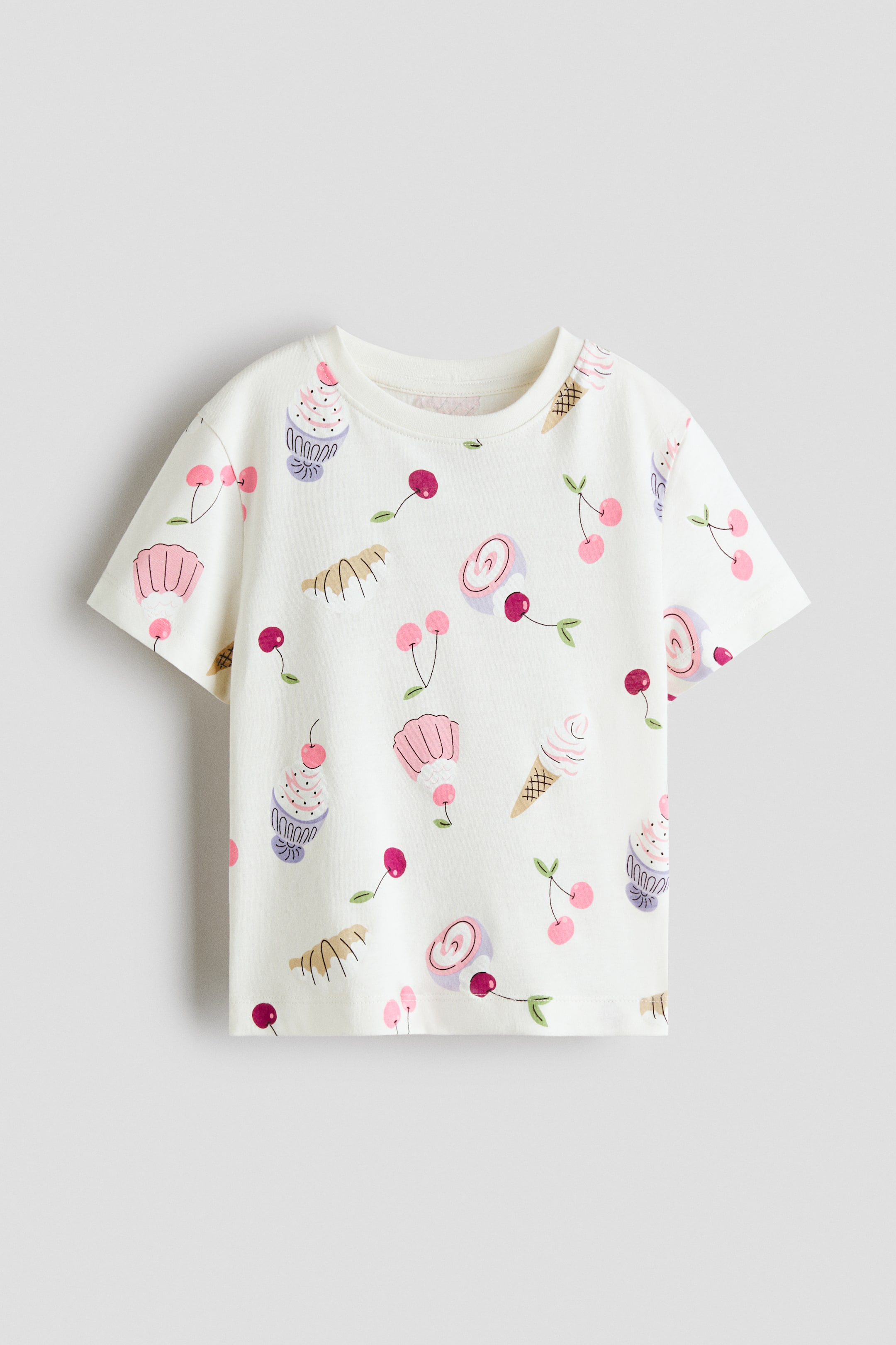 View larger image: A short-sleeved, off-white t-shirt featuring a crew neck showcases a vibrant pattern of colourful cupcakes, ice cream cones, lollipops, and cherries across its front.