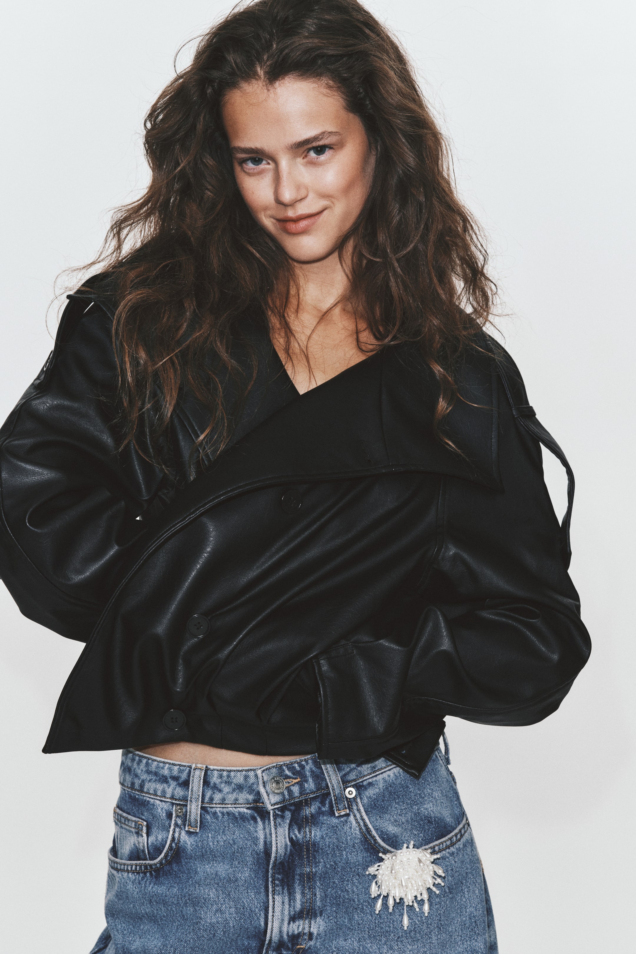 View larger image: An oversized black double-breasted jacket in a leather-like fabric is worn by the model, styled with blue denim jeans. The jacket features a wide draped collar, button fastenings, and long shoulder tabs extending down the raglan sleeves.