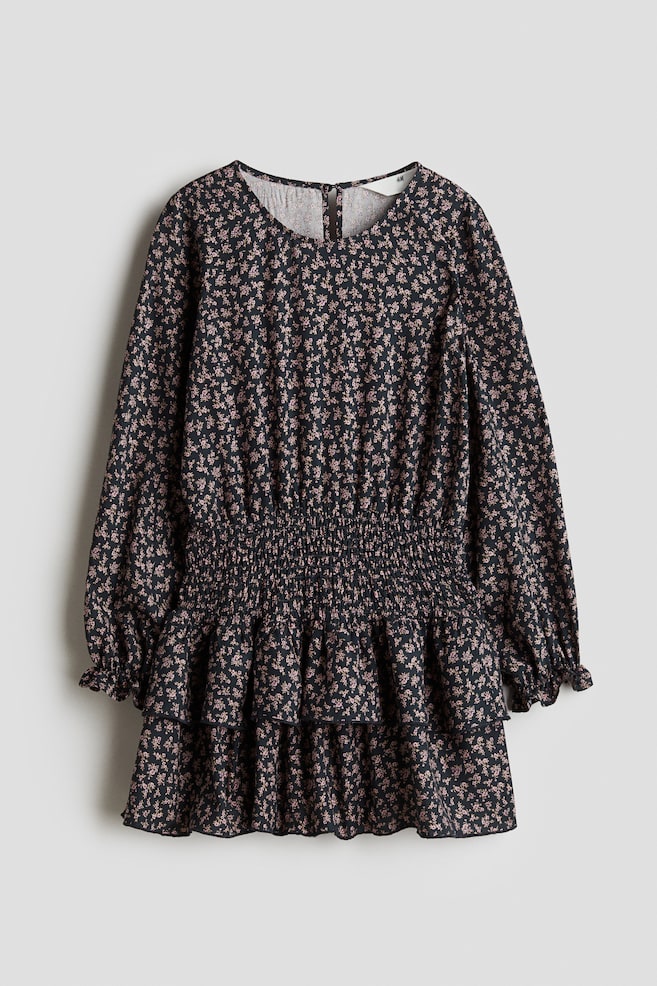 A dark dress with a small all-over floral pattern is displaying its front. It is featuring a round neckline, long sleeves with elasticated ruffled cuffs, a smocked elasticated waist, and a tiered skirt with two gathered ruffles. - 1