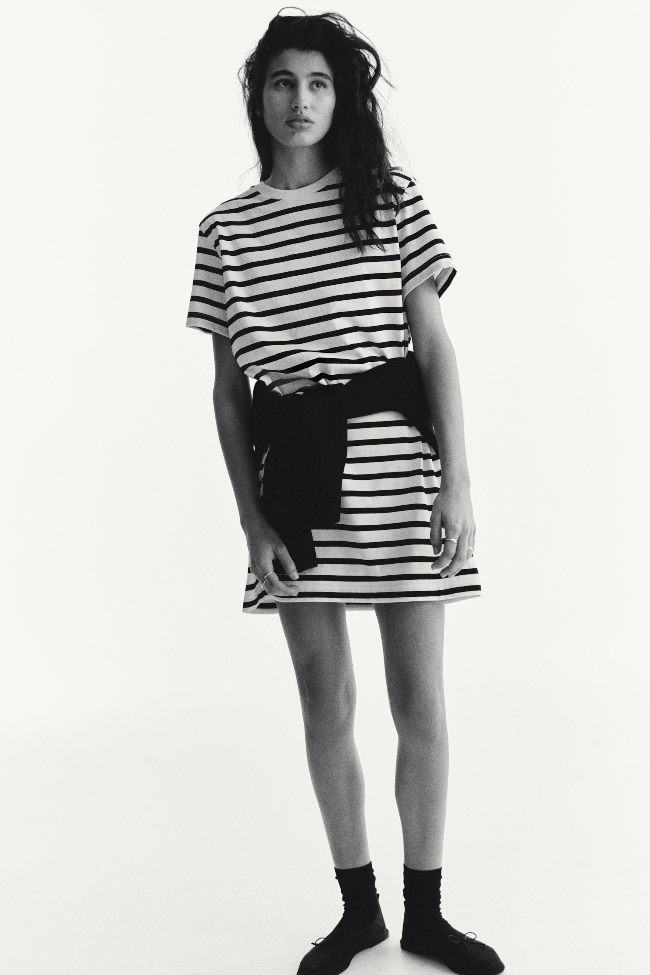 Xem ảnh lớn hơn: Chiếc váy áo thun sọc đen trắng này có tay ngắn, cổ tròn và chiều dài ngắn, hướng về phía trước. Một chiếc áo len đen được buộc quanh eo, tạo dáng thon gọn.