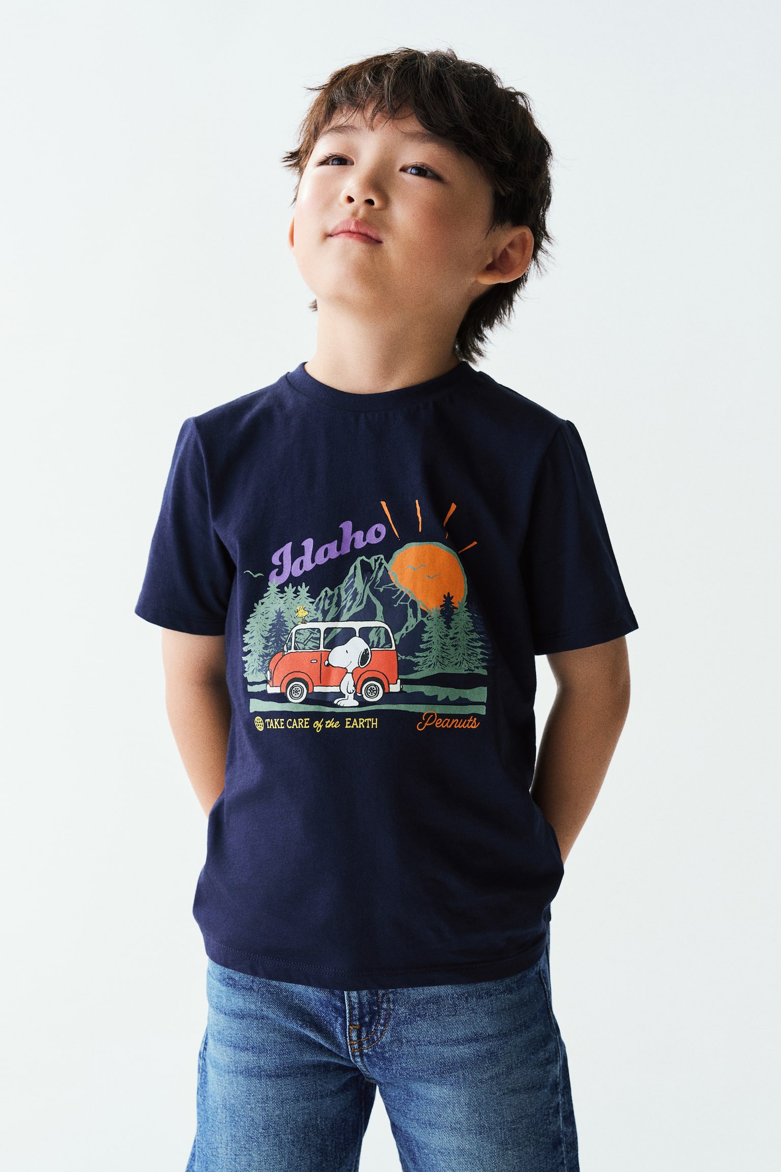 The model is wearing a navy blue cotton jersey T-shirt featuring a round, rib-trimmed neckline and a colourful graphic print of Snoopy in a mountain setting. The short-sleeved tee is paired with blue denim jeans. - 1