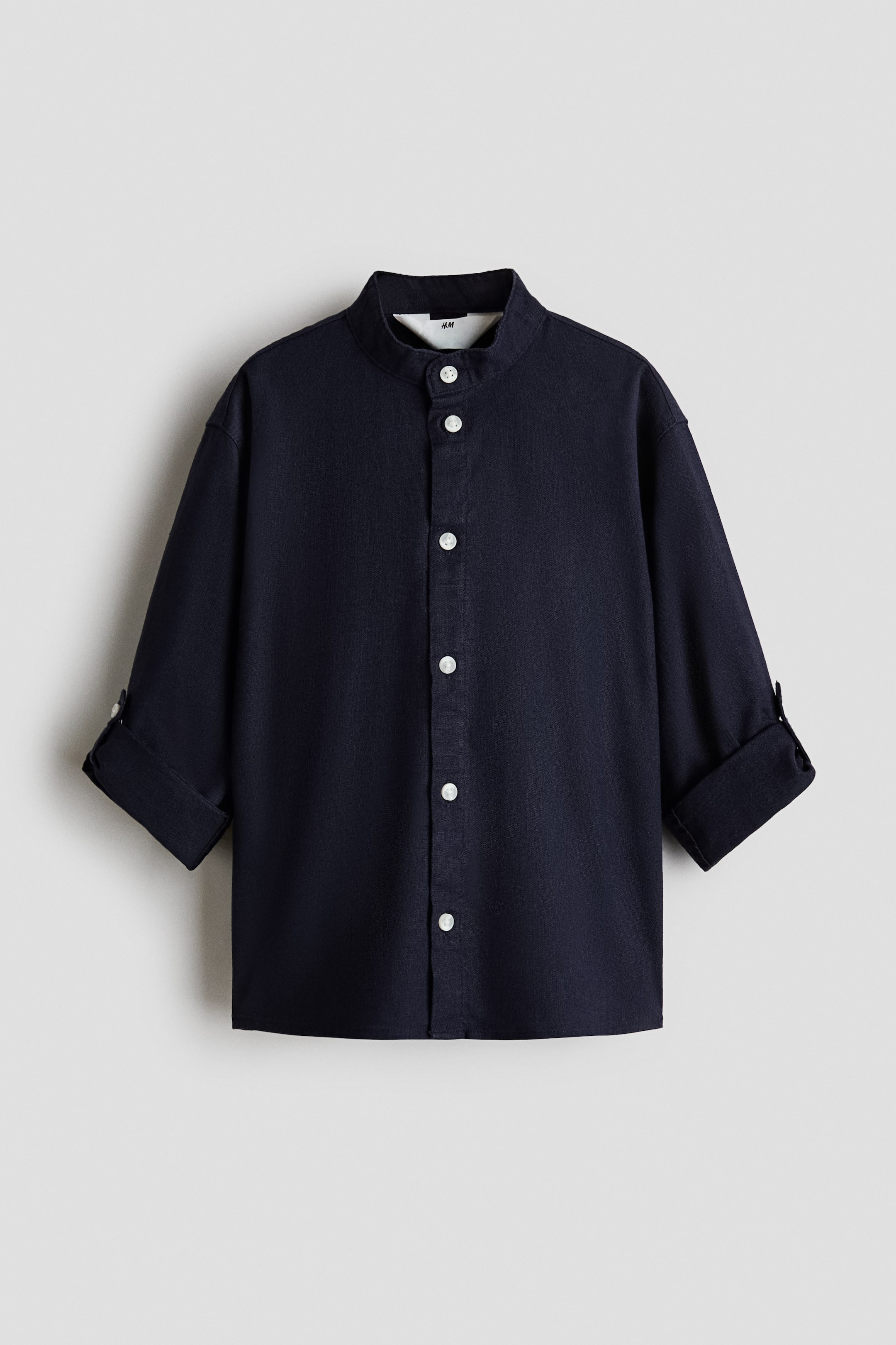 View larger image: A dark blue shirt is shown from the front, featuring a mandarin collar and a full placket with white buttons. Its long sleeves are rolled up to three-quarter length and secured with a buttoned strap.