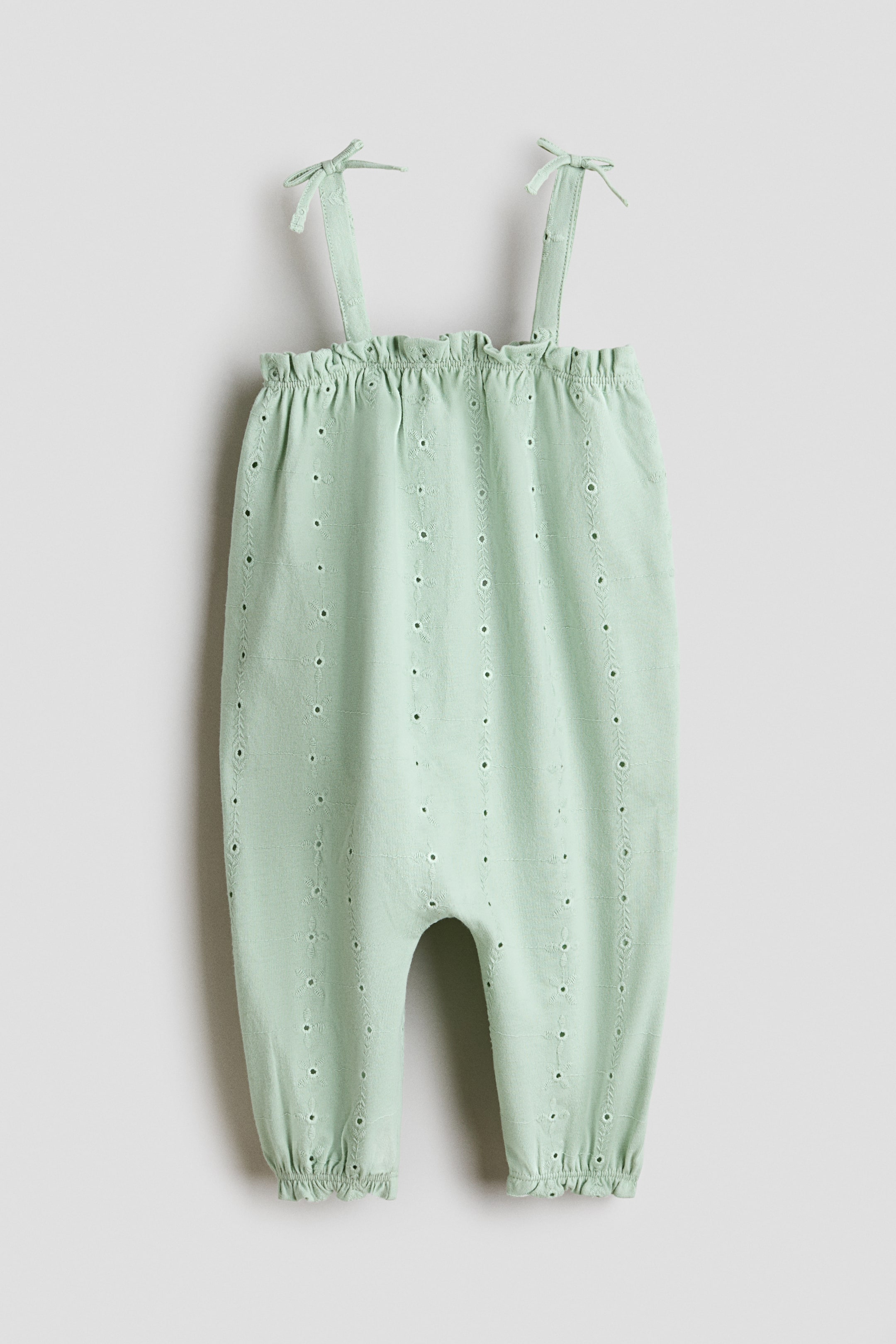 View larger image: A light green romper suit is showcased in front, crafted from soft cotton jersey with an all-over broderie anglaise pattern. It features a straight-cut top with shoulder straps tied into bows, and elasticated, frill-trimmed seams are visible around the top and at the leg hems.