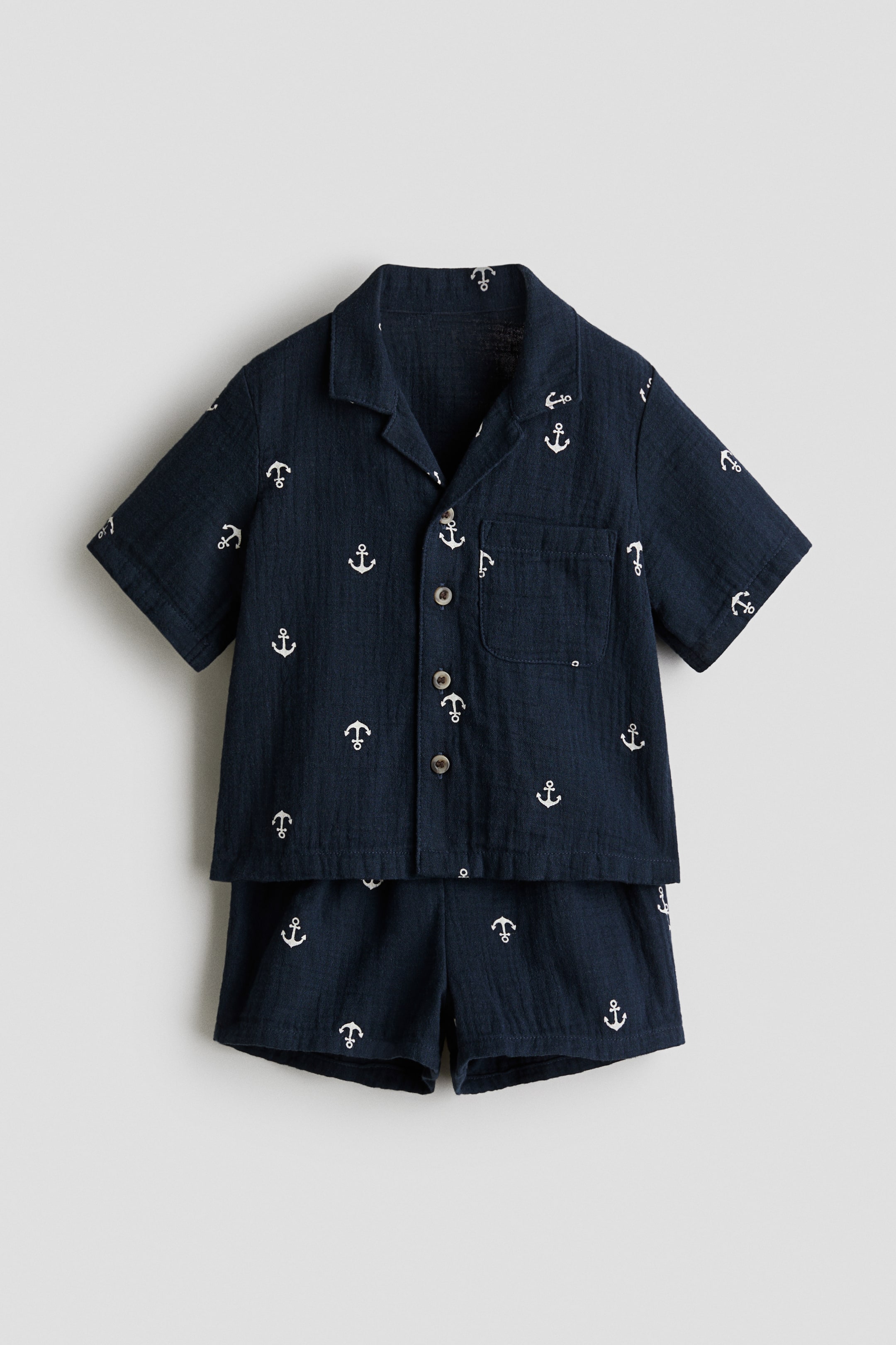 View larger image: A set of a shirt and shorts is presented FRONT, crafted from navy blue woven fabric adorned with a white anchor pattern. The straight-cut shirt features a resort collar, buttons down the front, short sleeves, and an open chest pocket, while the shorts boast an elasticated waist.