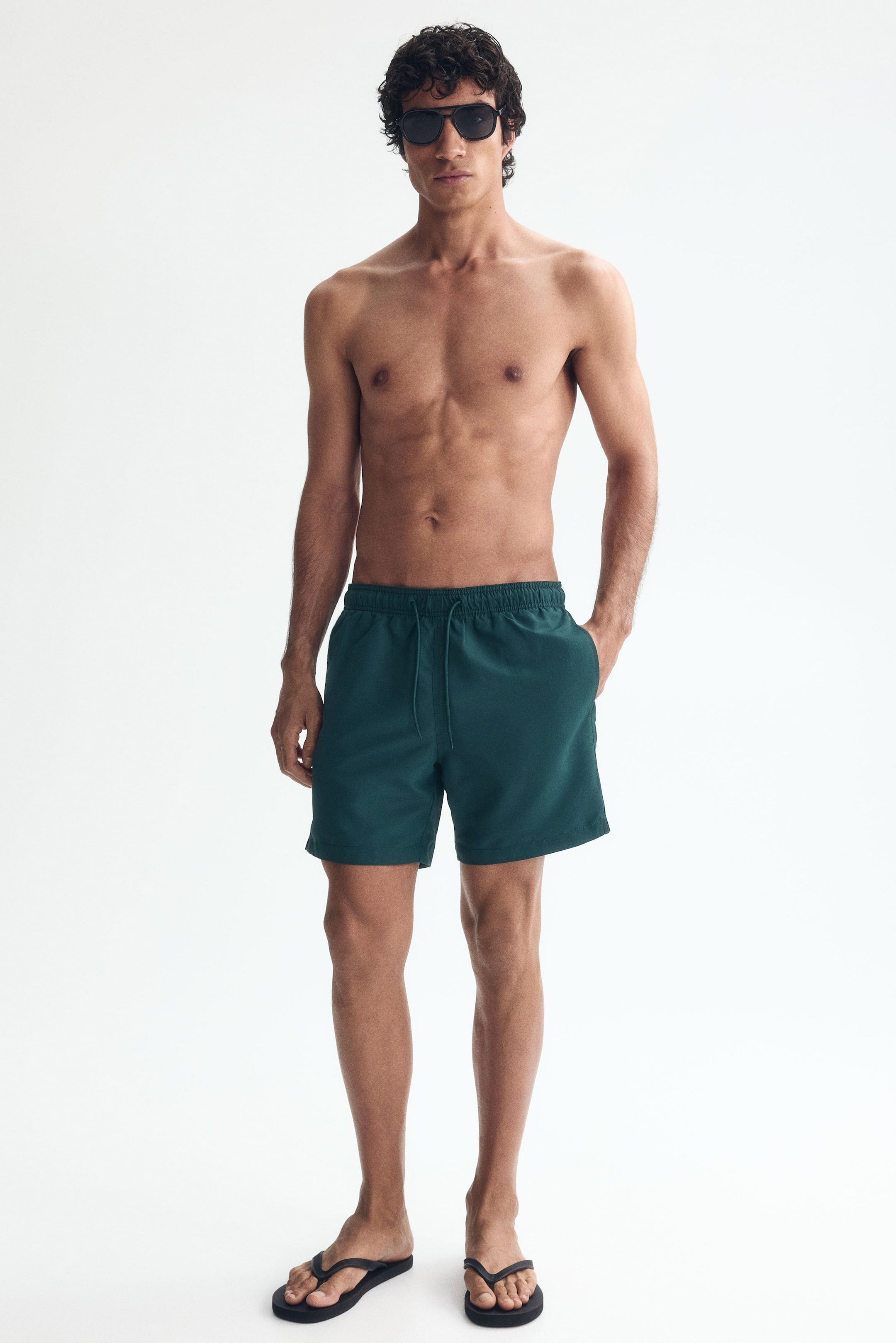 View larger image: The model is wearing dark green swim shorts made from woven fabric. These shorts have a covered elastic waistband with a drawstring, side pockets with the model's right hand in one, and a small slit on the side.