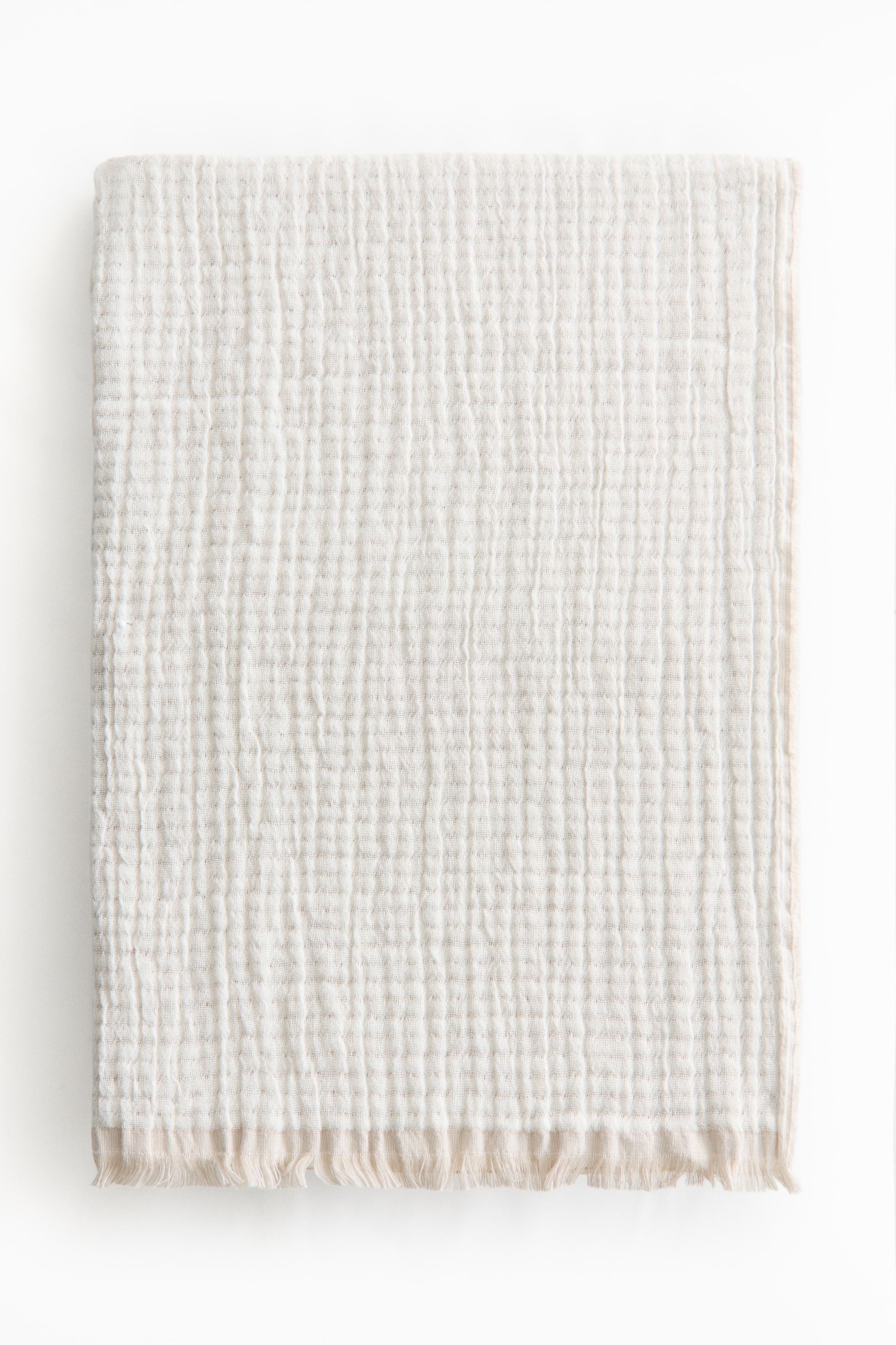 View larger image: The front of a light-coloured cotton muslin blanket displays a crinkled texture. Short fringes are visible along its bottom edge.