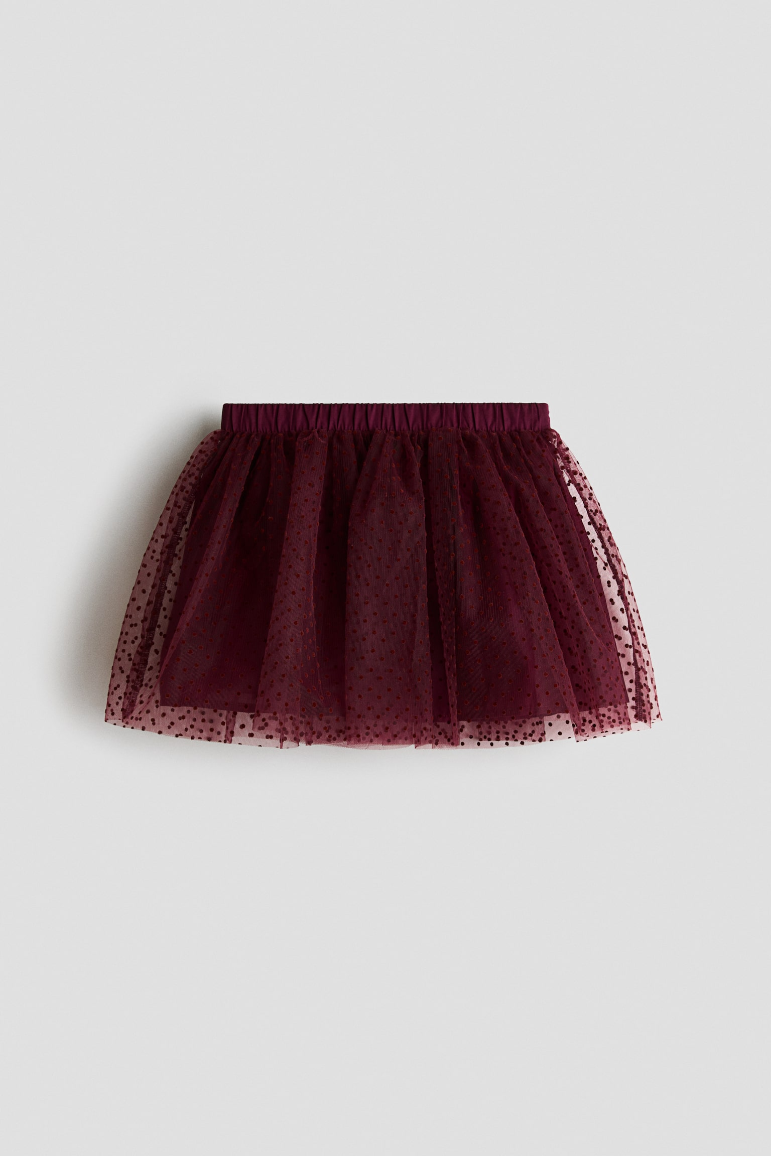 A dark red flared tulle skirt is shown from the front, featuring an all-over flocked polka dot print and an elasticated waistband. The sheer outer tulle layer is gathered at the waist and drapes over a solid, matching lining.