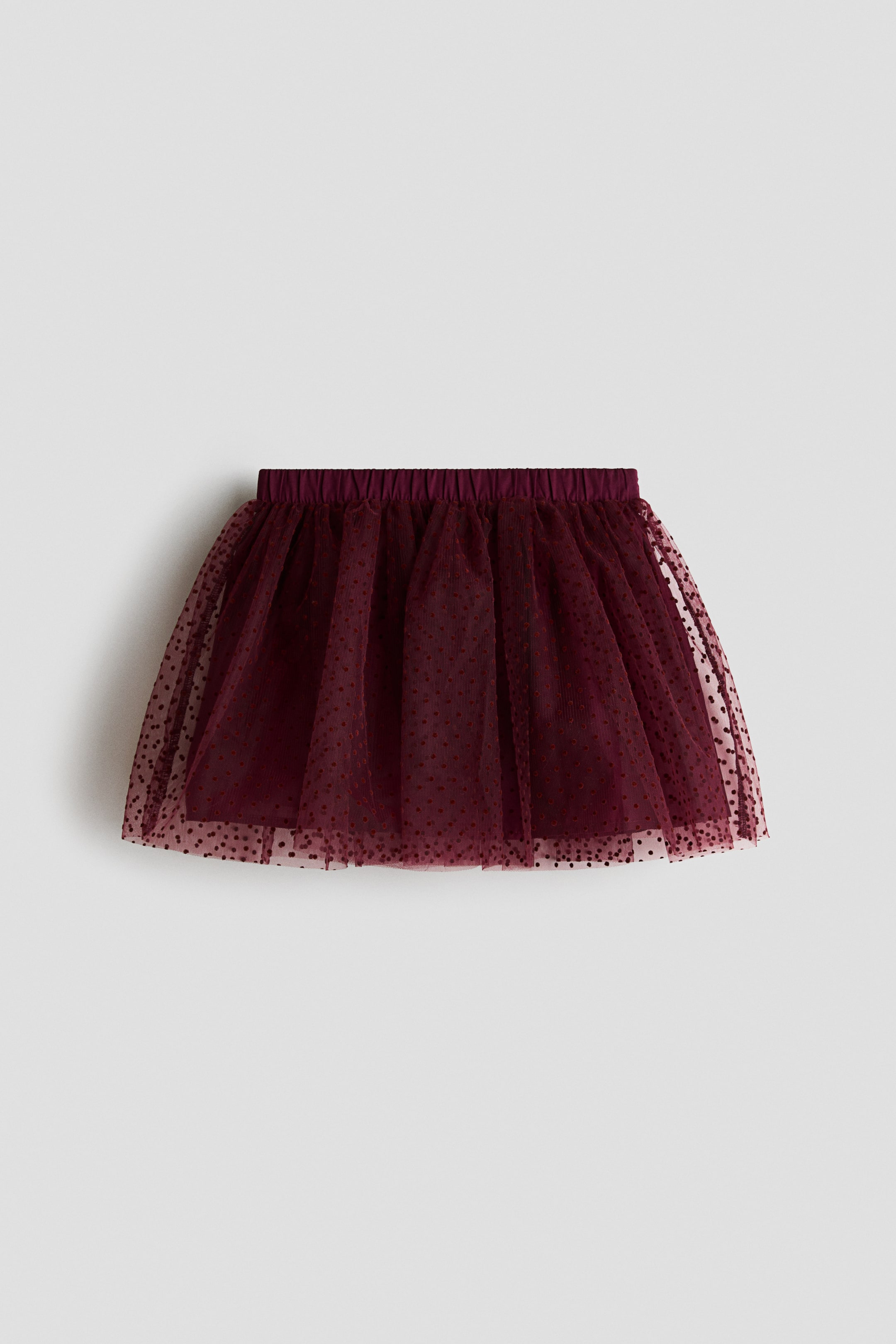 View larger image: A dark red flared tulle skirt is shown from the front, featuring an all-over flocked polka dot print and an elasticated waistband. The sheer outer tulle layer is gathered at the waist and drapes over a solid, matching lining.