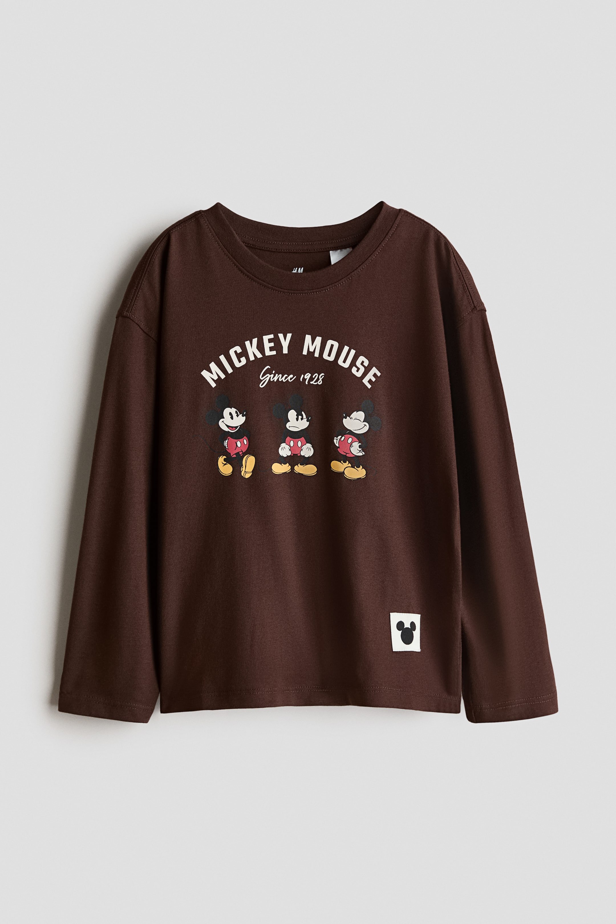 View larger image: A long-sleeved t-shirt in soft dark brown cotton jersey showcases a front print and a round, rib-trimmed neckline. The print features "MICKEY MOUSE Since 1928" in white curved text above three cartoon Mickey Mouse characters, and a small white square patch with a black Mickey Mouse silhouette is visible on the lower right.