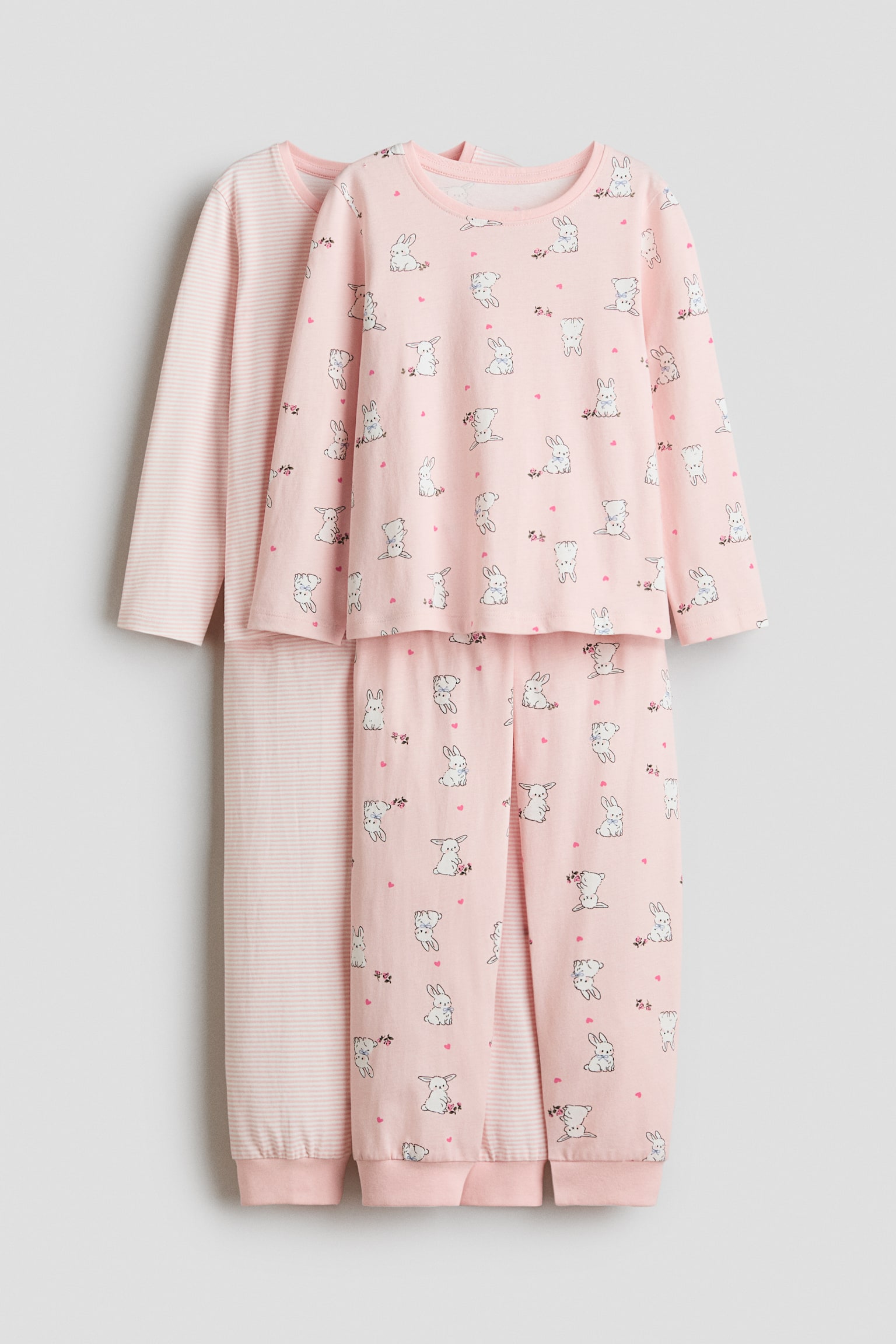 Two cotton jersey pyjama sets are hanging front-facing, with one set layered behind the other. The front set features an all-over print of white rabbits and small pink hearts on a light pink long-sleeve top and matching cuffed trousers. The partially visible set at the back features a light pink and white striped pattern on its matching long-sleeve top and trousers.