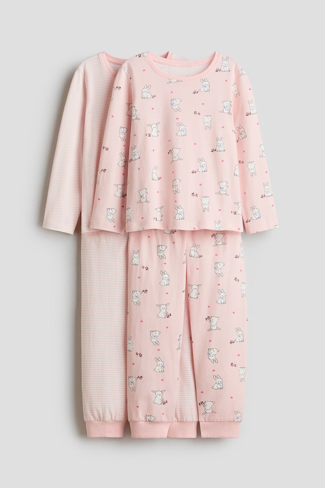 Two cotton jersey pyjama sets are hanging front-facing, with one set layered behind the other. The front set features an all-over print of white rabbits and small pink hearts on a light pink long-sleeve top with matching cuffed trousers. The partially visible set at the back features a light pink and white striped pattern on its matching long-sleeve top and trousers. - 1