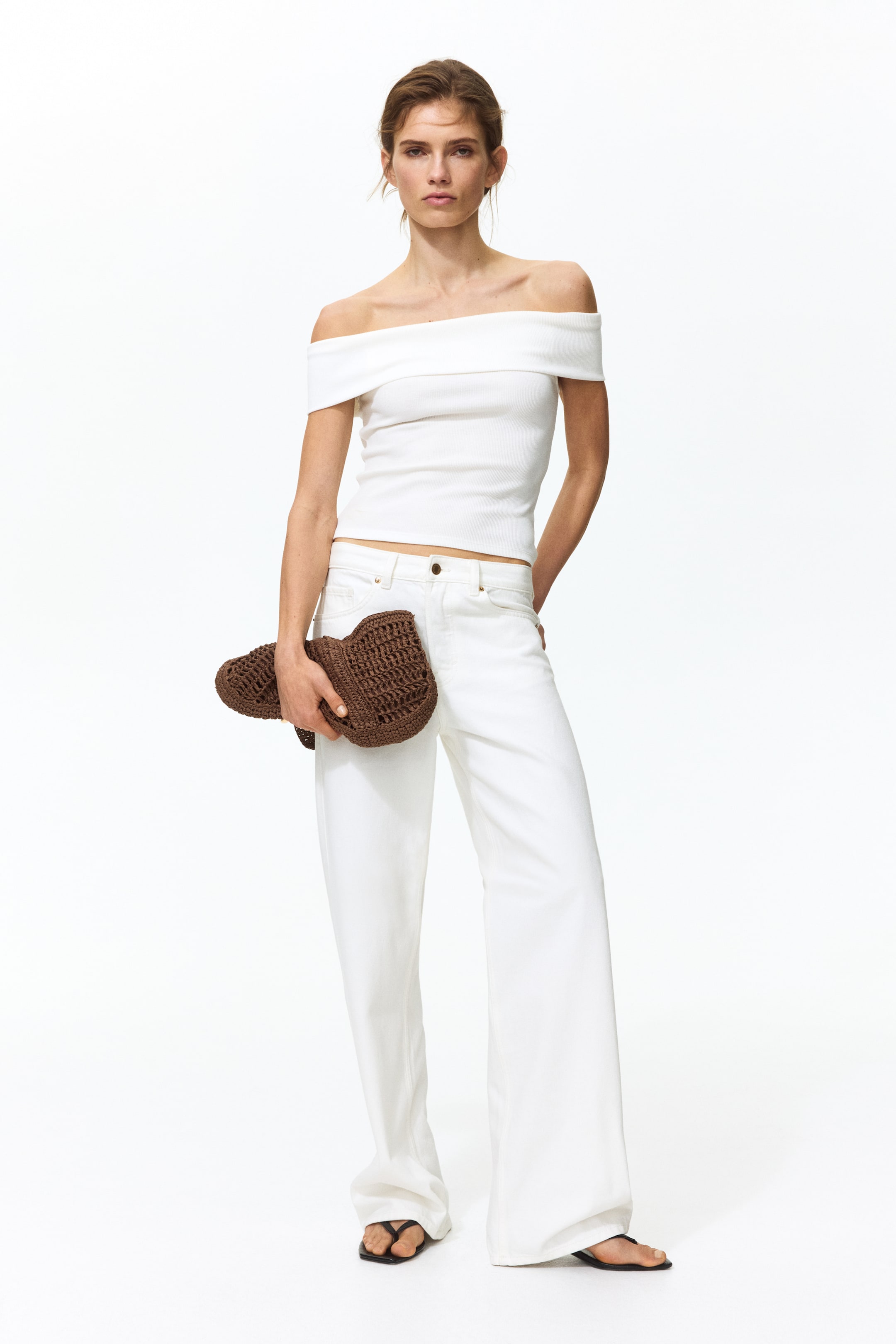 View larger image: A white off-the-shoulder ribbed knit top is showcased, featuring a wide folded neckline and short sleeves. This top is styled with white high-waisted wide-leg jeans, and the ensemble is accessorized with a brown crocheted clutch and black thong sandals.