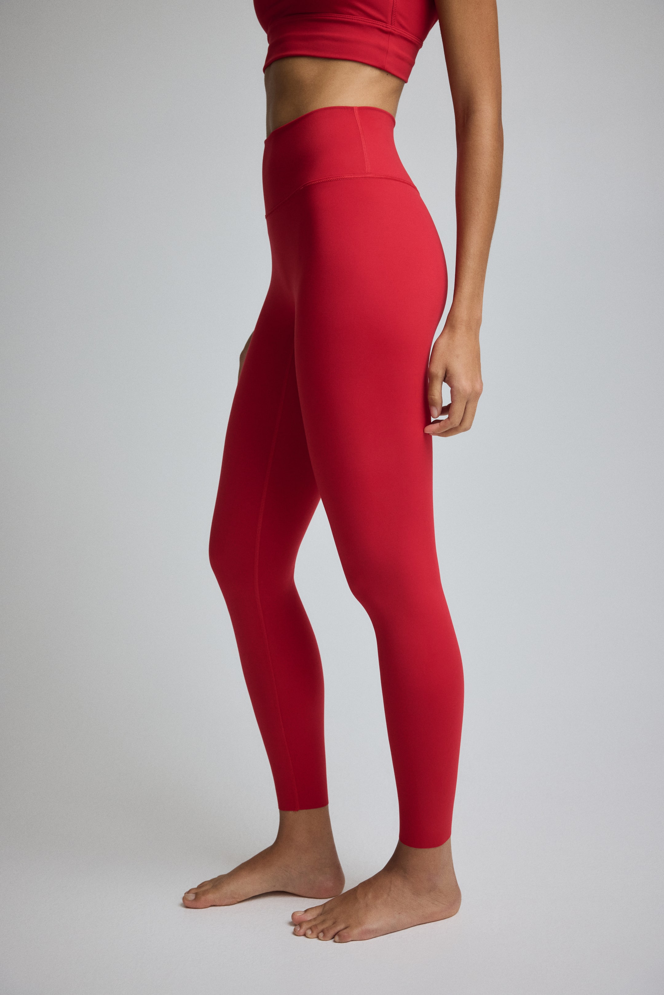 View larger image: Red leggings are being shown from a side view, featuring a high-waisted fit and a smooth, form-fitting fabric. They are extending to the ankles and are being paired with a matching red sports bra.