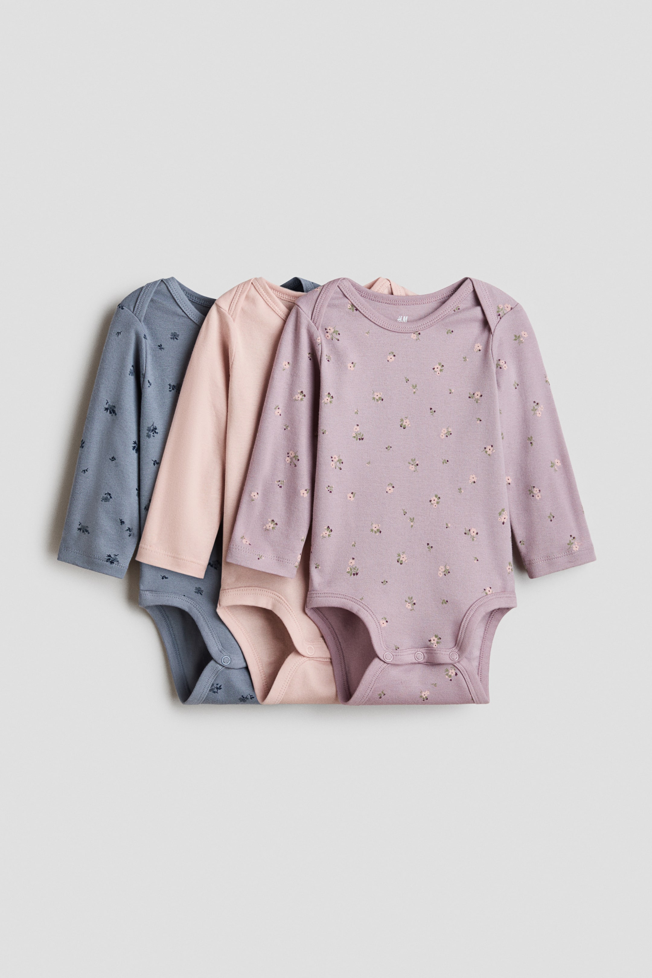 View larger image: Three long-sleeved soft cotton jersey bodysuits are shown front-facing, featuring an envelope neck and press-studs at the bottom. The front bodysuit is purple with a small floral print, a plain pink bodysuit is layered partially behind it, and a blue bodysuit with a subtle print is layered partially behind the pink.
