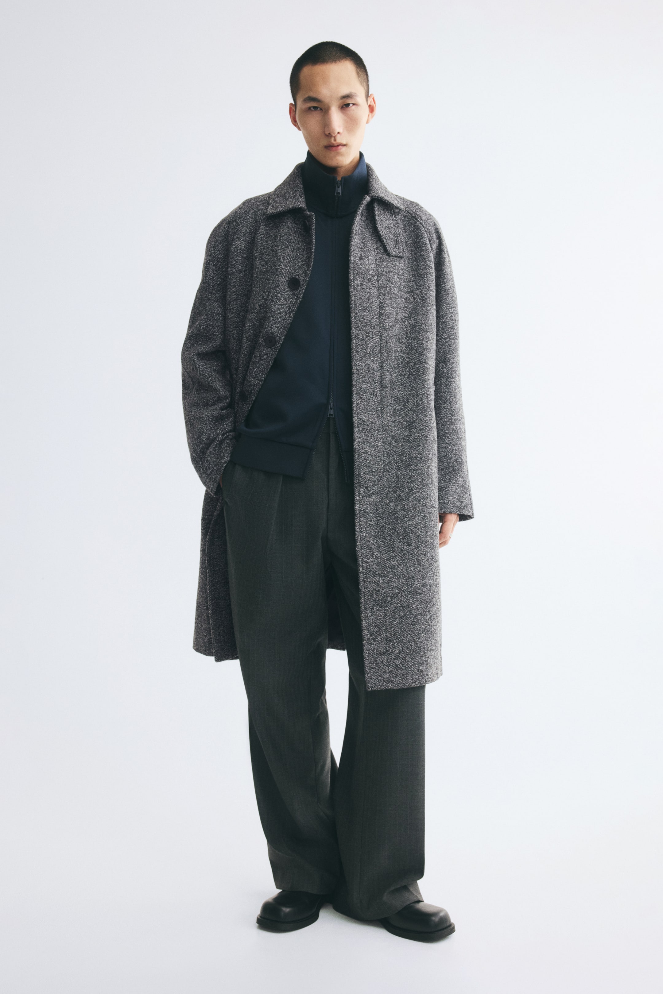 View larger image: A knee-length car coat in a grey, herringbone-weave wool blend is worn by the model with one hand resting in the side pocket. The coat is styled open, showcasing its classic collar and raglan sleeves, layered over a dark blue zip-up jacket and charcoal grey trousers.