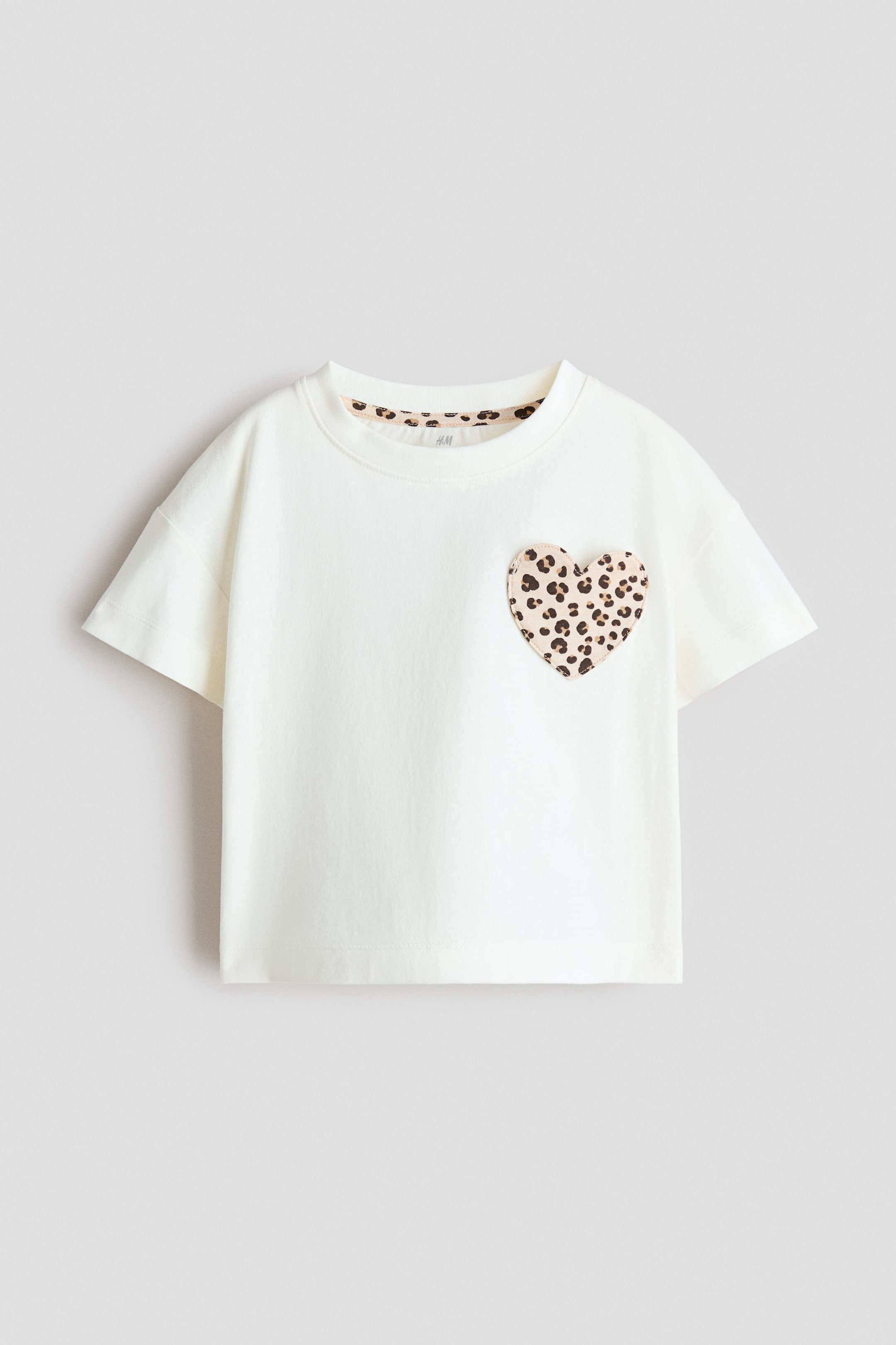 Ampliar la imagen: Una camiseta de color crema muestra su PARTE DELANTERA, con cuello redondo y mangas cortas. Un parche en forma de corazón con un estampado de leopardo es visible en el pecho izquierdo, y un ribete a juego con estampado de leopardo es visible a lo largo del interior del escote.