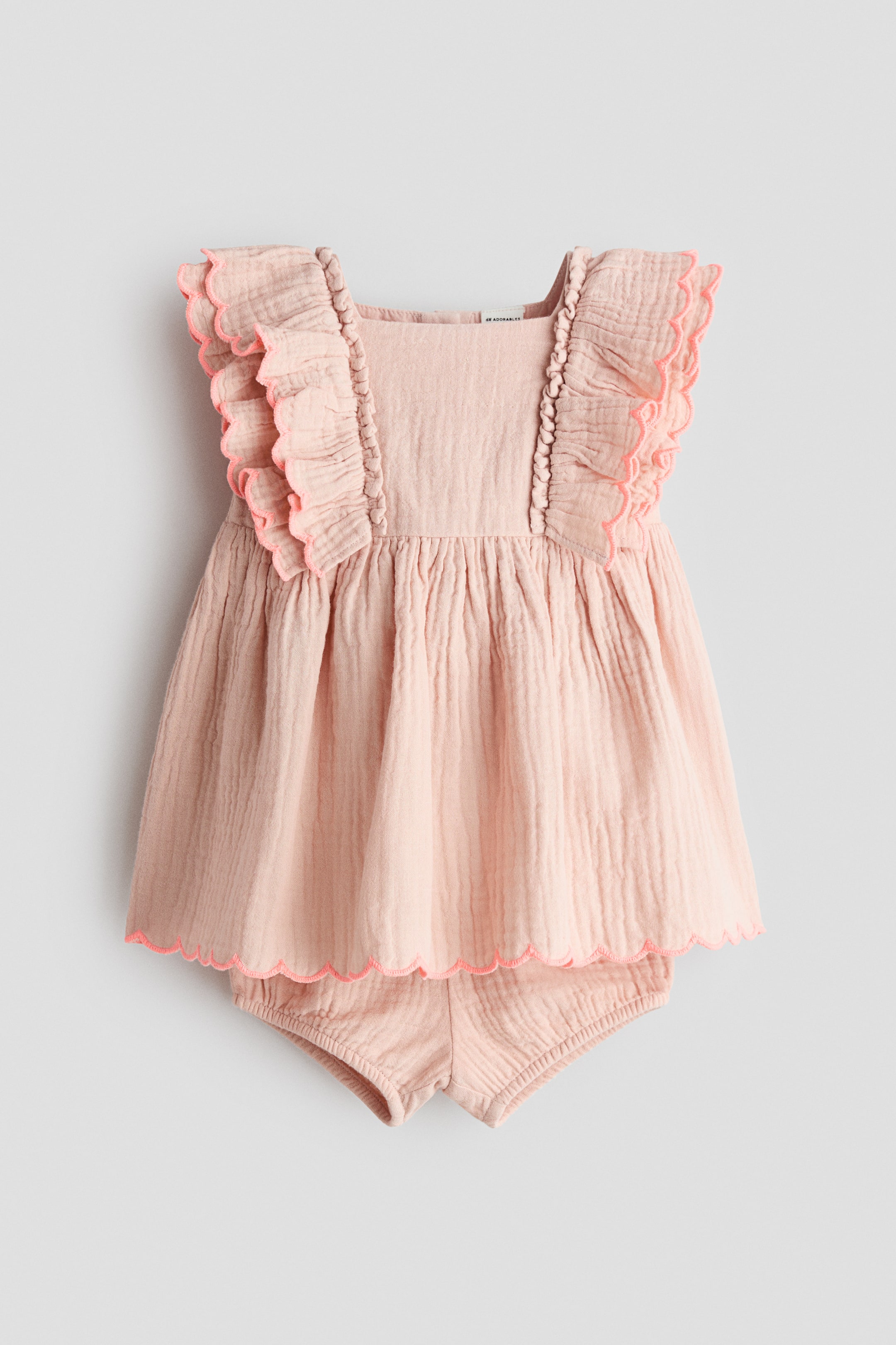 View larger image: The H&M Adorables set features a light pink dress and matching bloomers, both crafted from crinkled muslin fabric. The dress showcases a square neckline with a narrow yoke and double-layered scalloped frills, trimmed in a brighter pink, adorning the shoulders. Gathers form below the yoke, leading to a flared skirt with a scalloped hem, also trimmed in brighter pink. The matching bloomers are visible underneath, featuring elasticated leg openings.