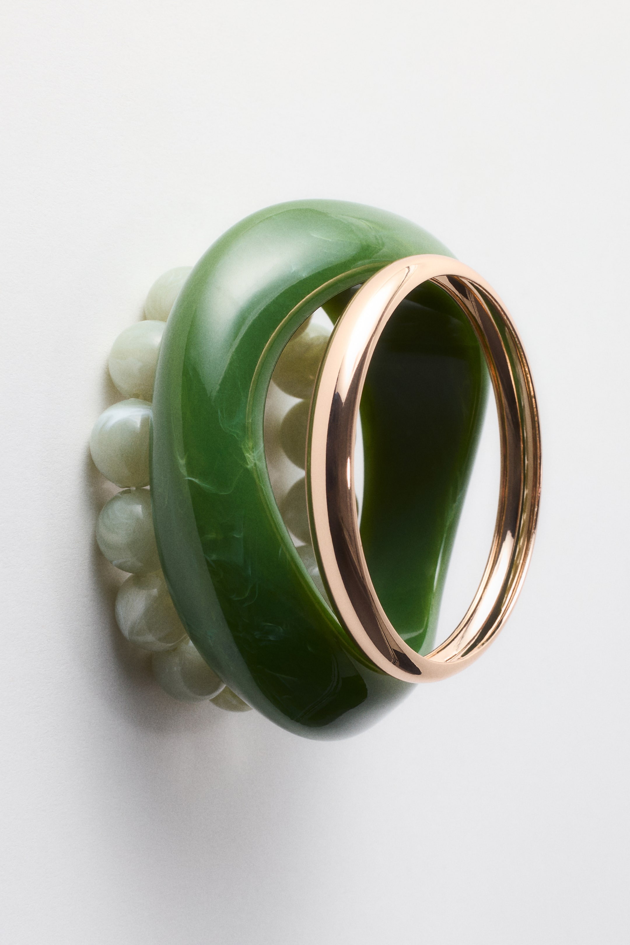 Grotere afbeelding bekijken: Een dikke, donkergroene gemarmerde armband staat rechtop op zijn rand, met een dunne, glanzende, roségoudkleurige armband gepositioneerd binnenin de opening. Een lichtgroene kralenarmband is gedeeltelijk zichtbaar onder de donkergroene armband.
