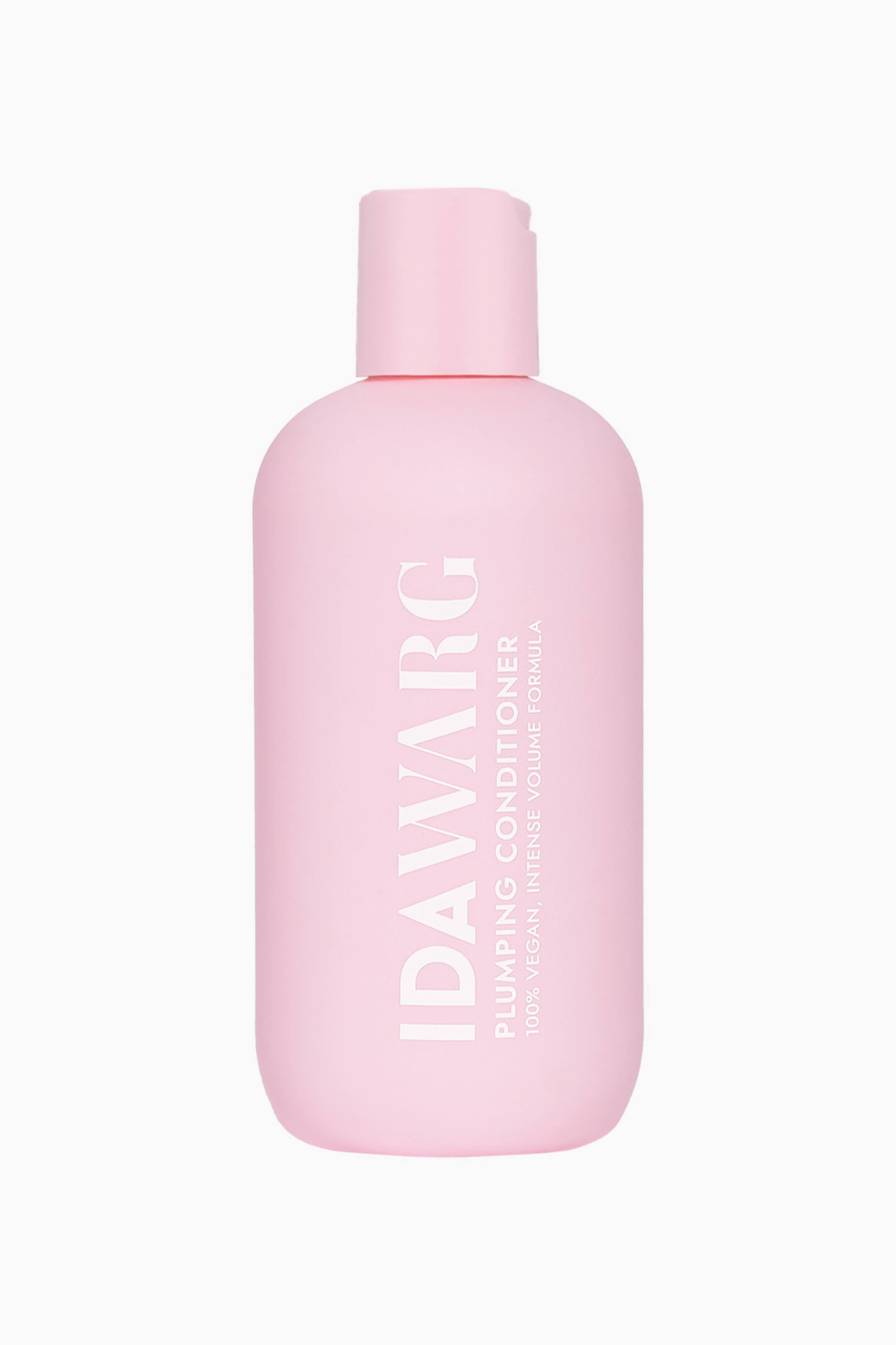 Vis større bilde: En matt rosa balsamflaske står oppreist, vendt fremover, og har en matchende rosa flip-kork. Hvit tekst merker forsiden, med "IDA WARG" vertikalt og "PLUMPING CONDITIONER" horisontalt.