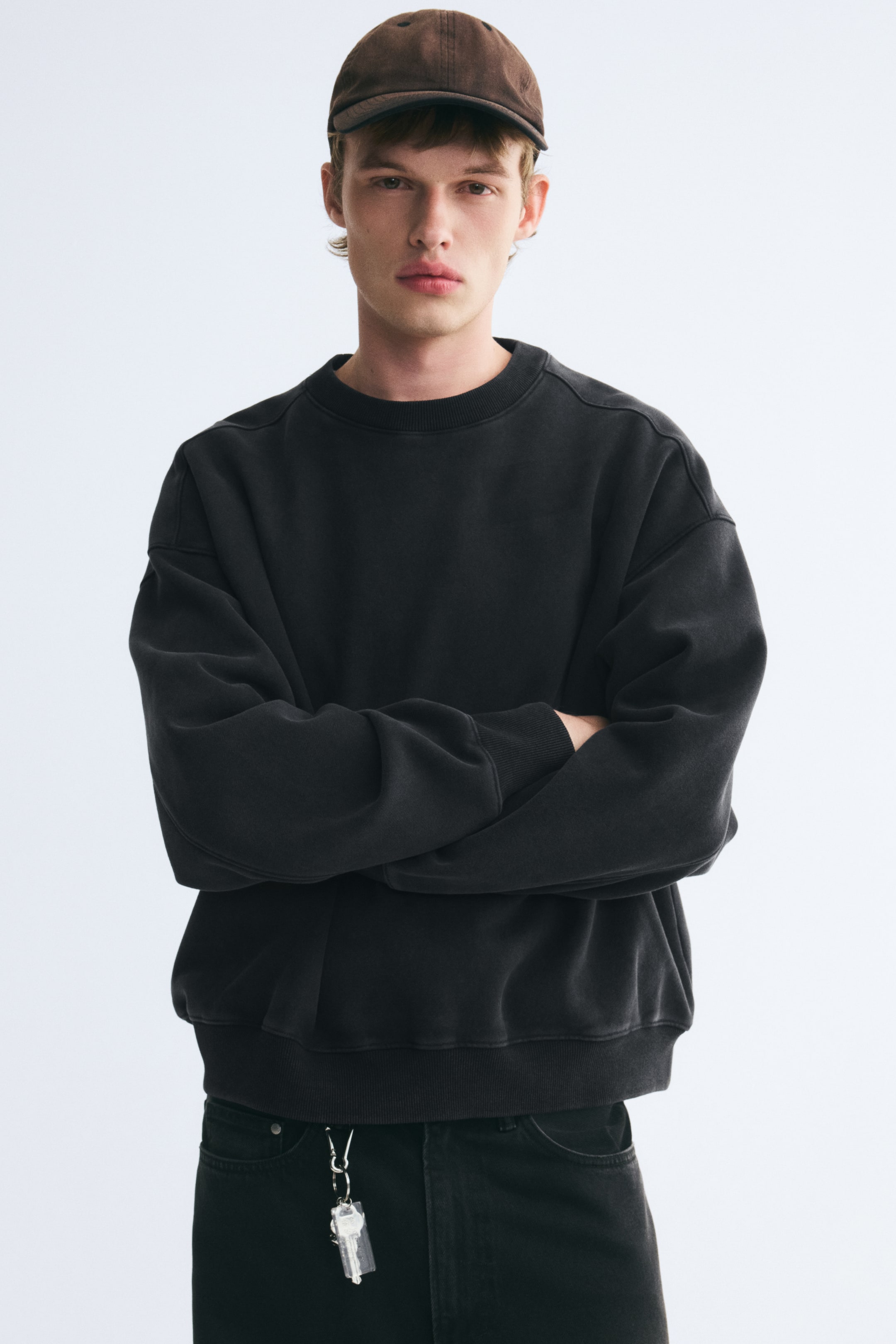 Преглед на по-голямо изображение: Моделът е облечен в oversized тъмносив суитшърт с рипсено обло деколте, спуснати рамене и рипсени маншети и подгъв. Моделът е в изправена поза със скръстени ръце, подчертавайки свободния, широк силует на дрехата.