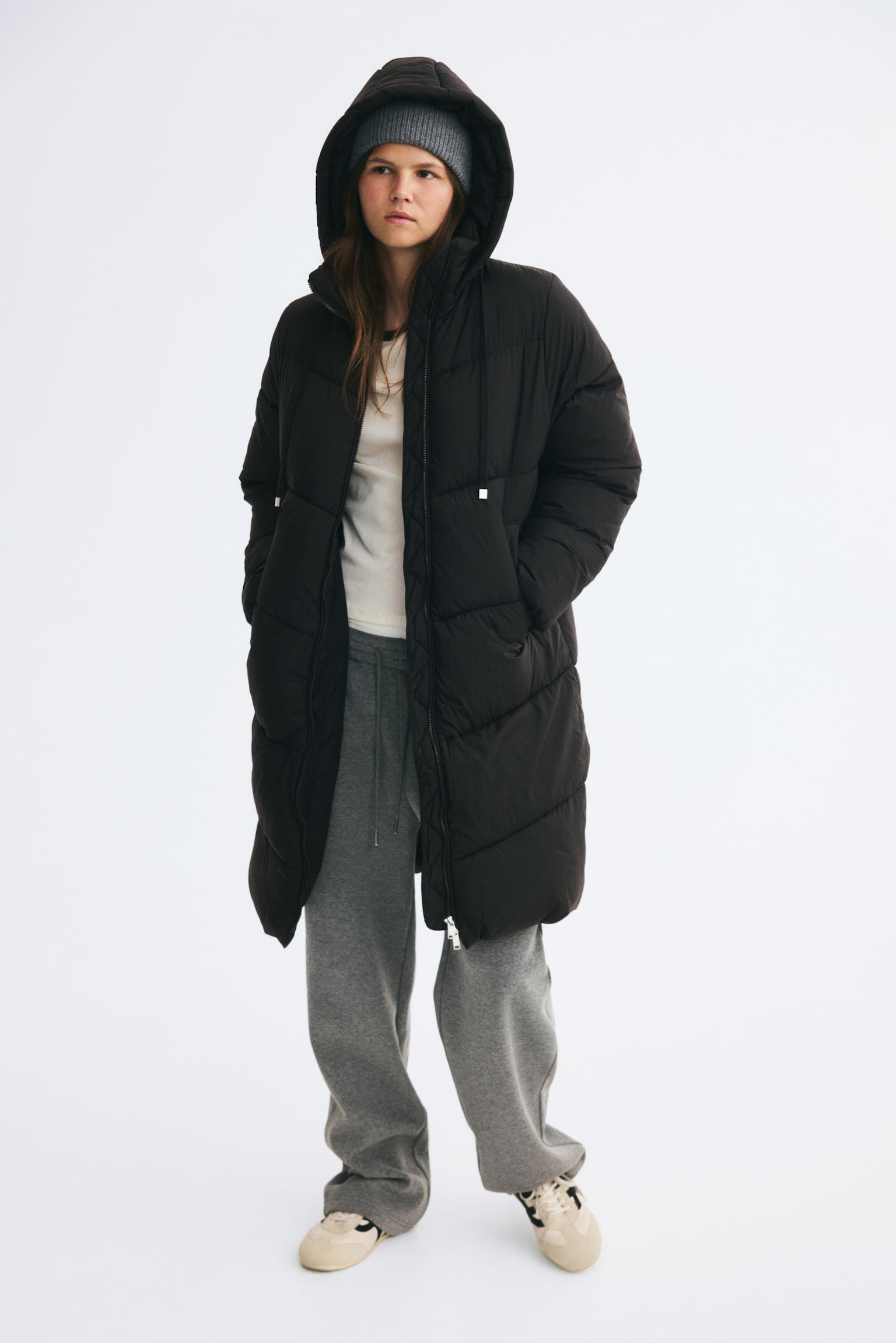 An oversized, knee-length black puffer coat in quilted nylon is worn by the model with the drawstring hood up over a grey knit beanie. The partially unzipped coat reveals a white T-shirt and grey sweatpants, while the model is standing with their hands placed in the front welt pockets. - 1