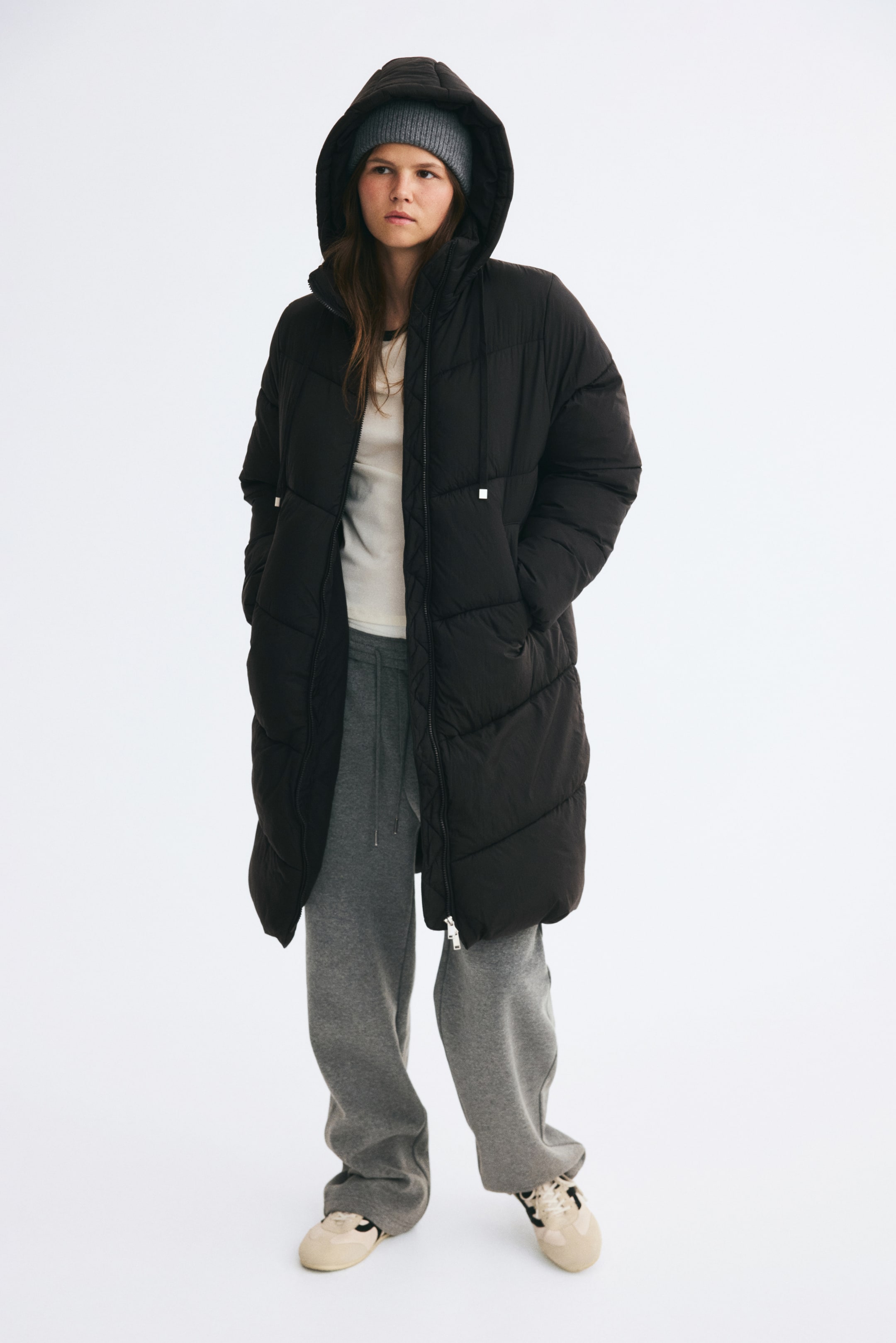 Agrandir l'image: Un manteau matelassé noir oversize, mi-long jusqu’aux genoux, en nylon matelassé, est porté par le mannequin, avec la capuche à cordon portée sur un bonnet en maille gris. Le manteau, partiellement ouvert, révèle un t-shirt blanc et un pantalon de jogging gris, tandis que le mannequin se tient debout, les mains glissées dans les poches passepoilées avant.
