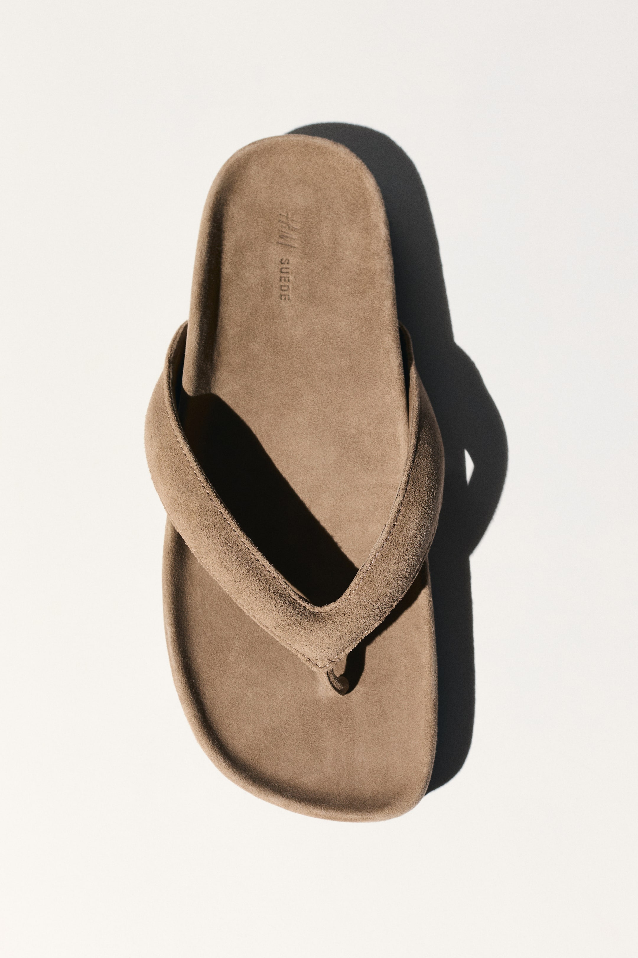 View larger image: A light brown suede thong sandal is positioned vertically, showcasing its upper surface. It features wide suede straps and a matching suede footbed with the text "H&M SUEDE" embossed near the top.