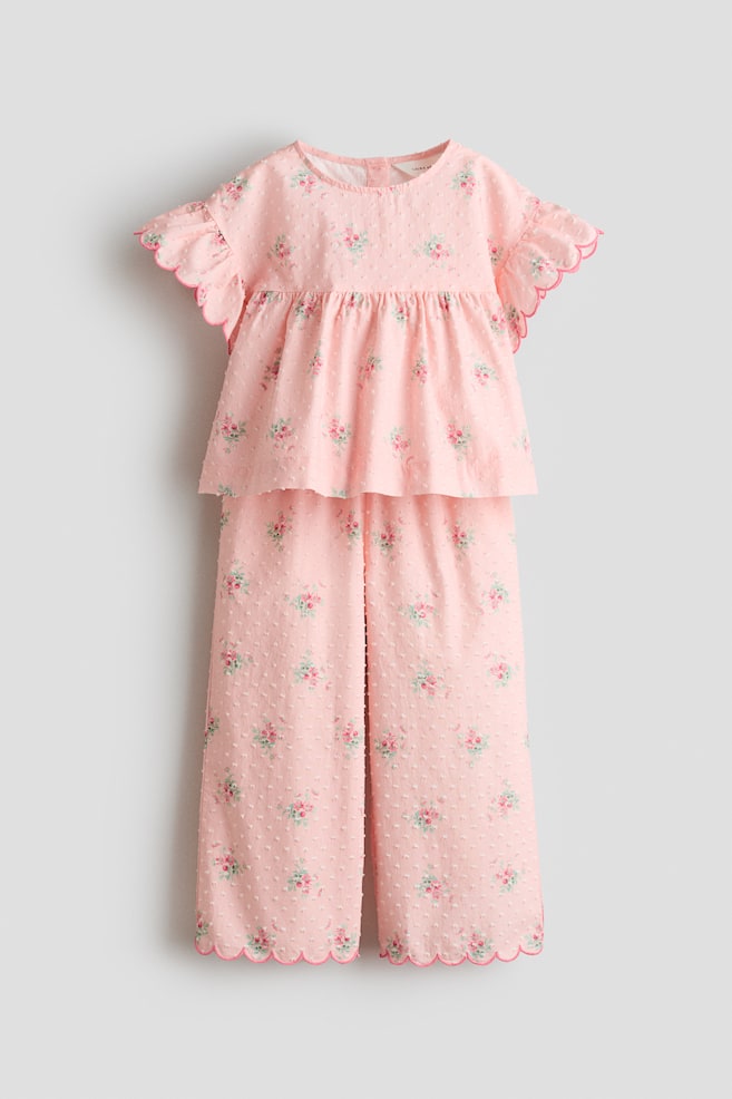 A pink two-piece set is presented from the front, featuring a top with a round neckline, short flutter sleeves with scalloped edges, and a gathered waist. The matching wide-leg trousers showcase scalloped hems, while both pieces display an all-over raised dot texture and a repeating floral pattern of small roses. - 1