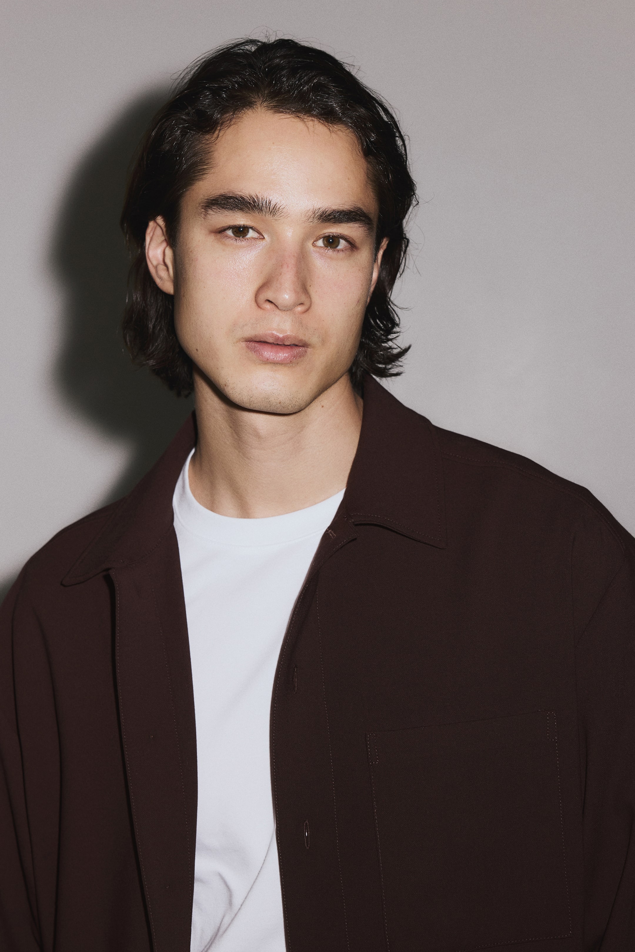 Ampliar la imagen: Una camisa de botones color marrón oscuro se lleva abierta sobre una camiseta blanca de cuello redondo. La camisa presenta un cuello amplio y un único bolsillo parche en el pecho izquierdo.