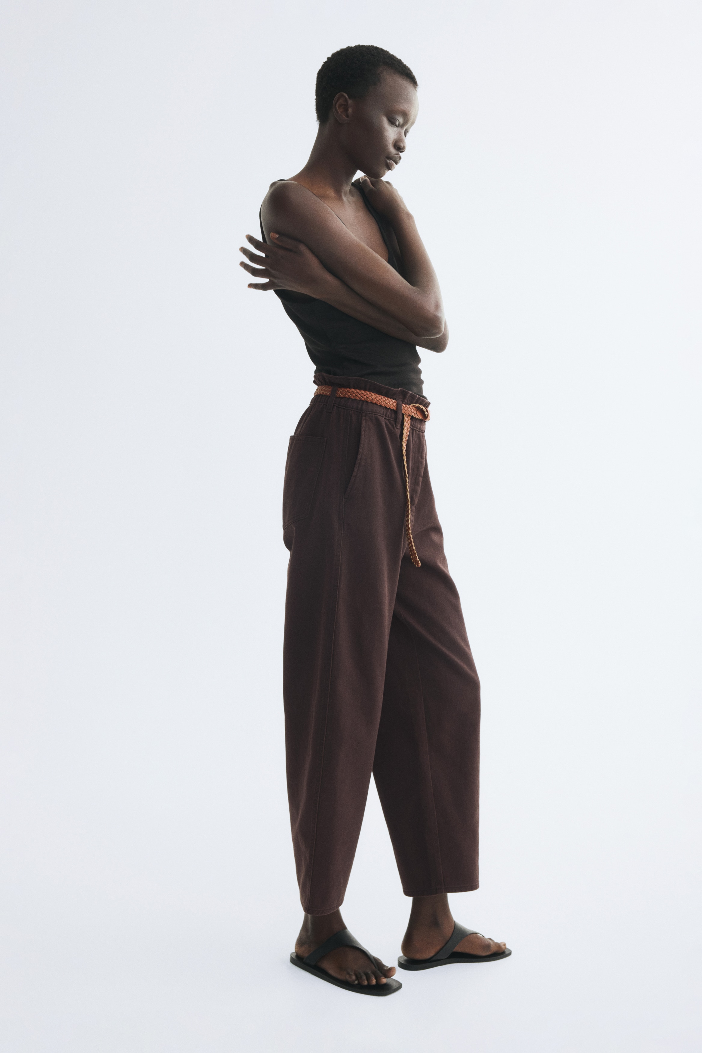 Die dunkelbraune Hose wird von vorne präsentiert und verfügt über eine geraffte hohe Taille mit einem braunen geflochtenen Gürtel, der durch die Schlaufen gezogen und mit einer silberfarbenen Schnalle gesichert ist. Seitentaschen sind sichtbar, und die Hose ist mit einem weit geschnittenen, verkürzten Silhouette-Design gestaltet.