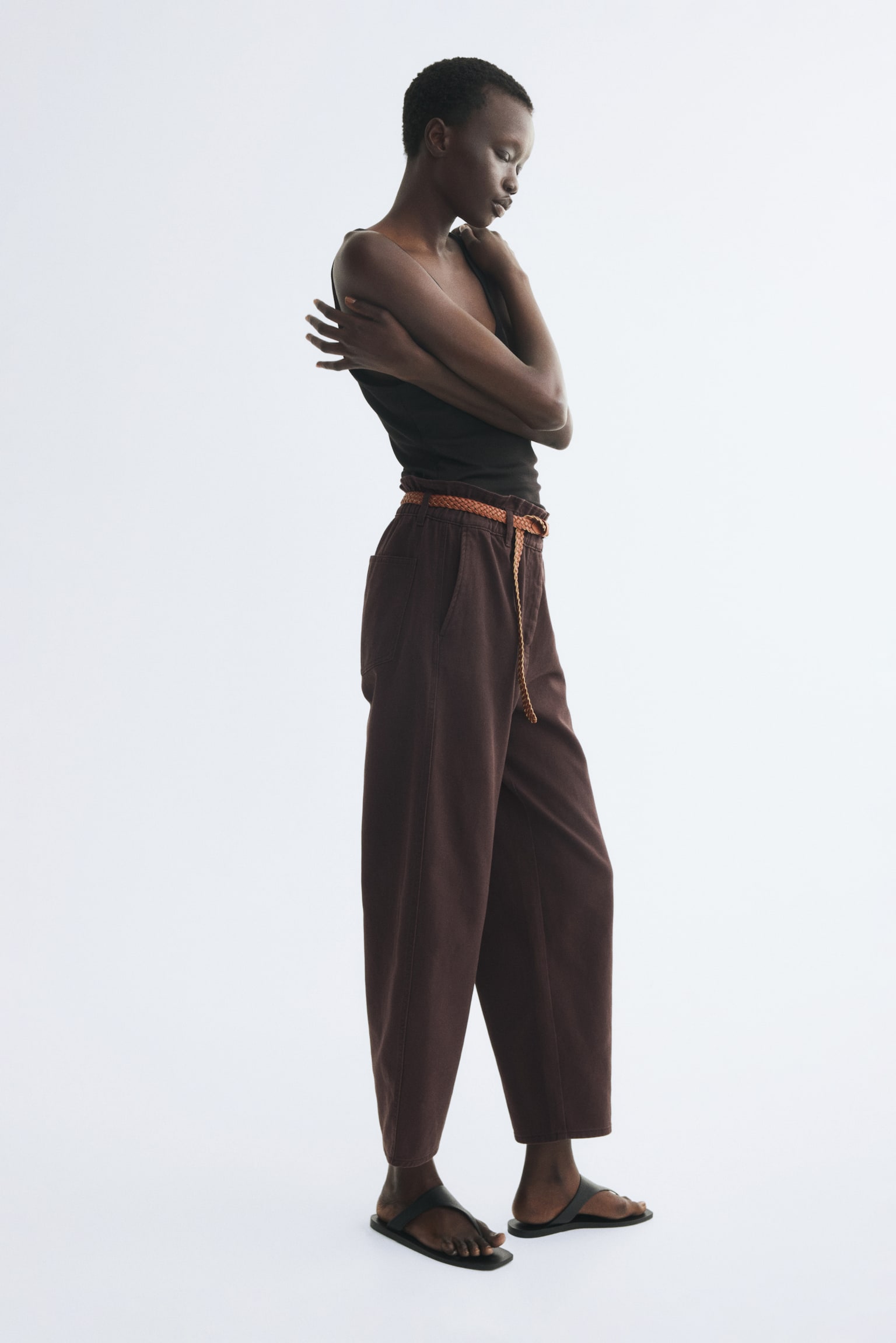 The dark brown trousers are showcased from the front, boasting a gathered high waist with a brown braided belt threaded through its loops and fastened by a silver-toned buckle. Side pockets are visible, and the trousers are crafted with a wide-leg, cropped silhouette. - 1