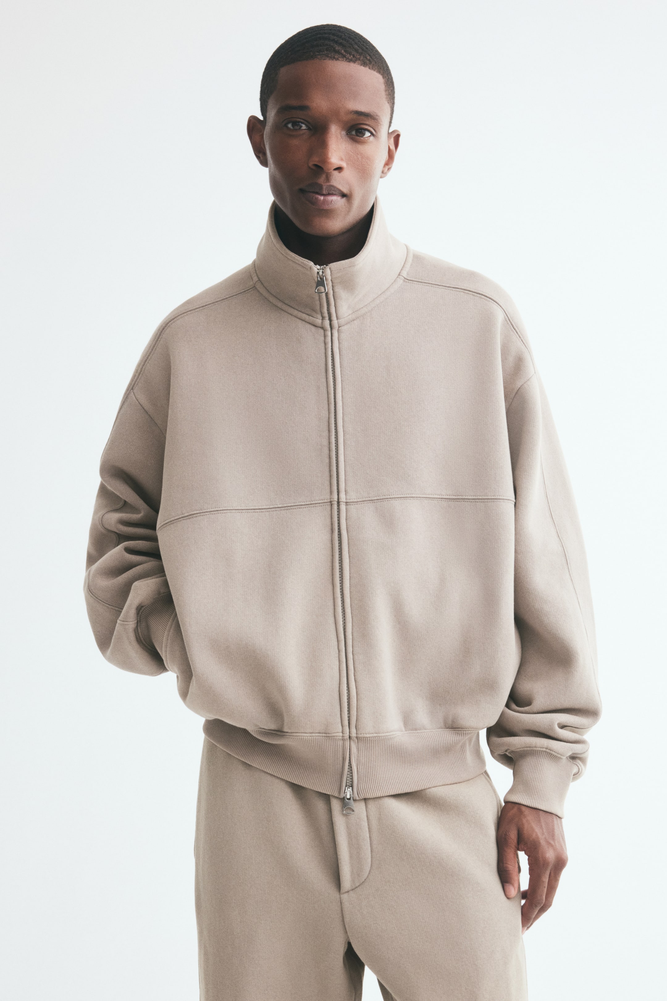 Voir une image plus grande: Une veste de survêtement beige à coupe oversize est portée par le mannequin, qui a une main glissée dans une poche latérale discrète. La veste présente un col montant, une fermeture éclair avant à double sens et des finitions côtelées aux poignets et à l’ourlet, agencée avec un pantalon assorti.