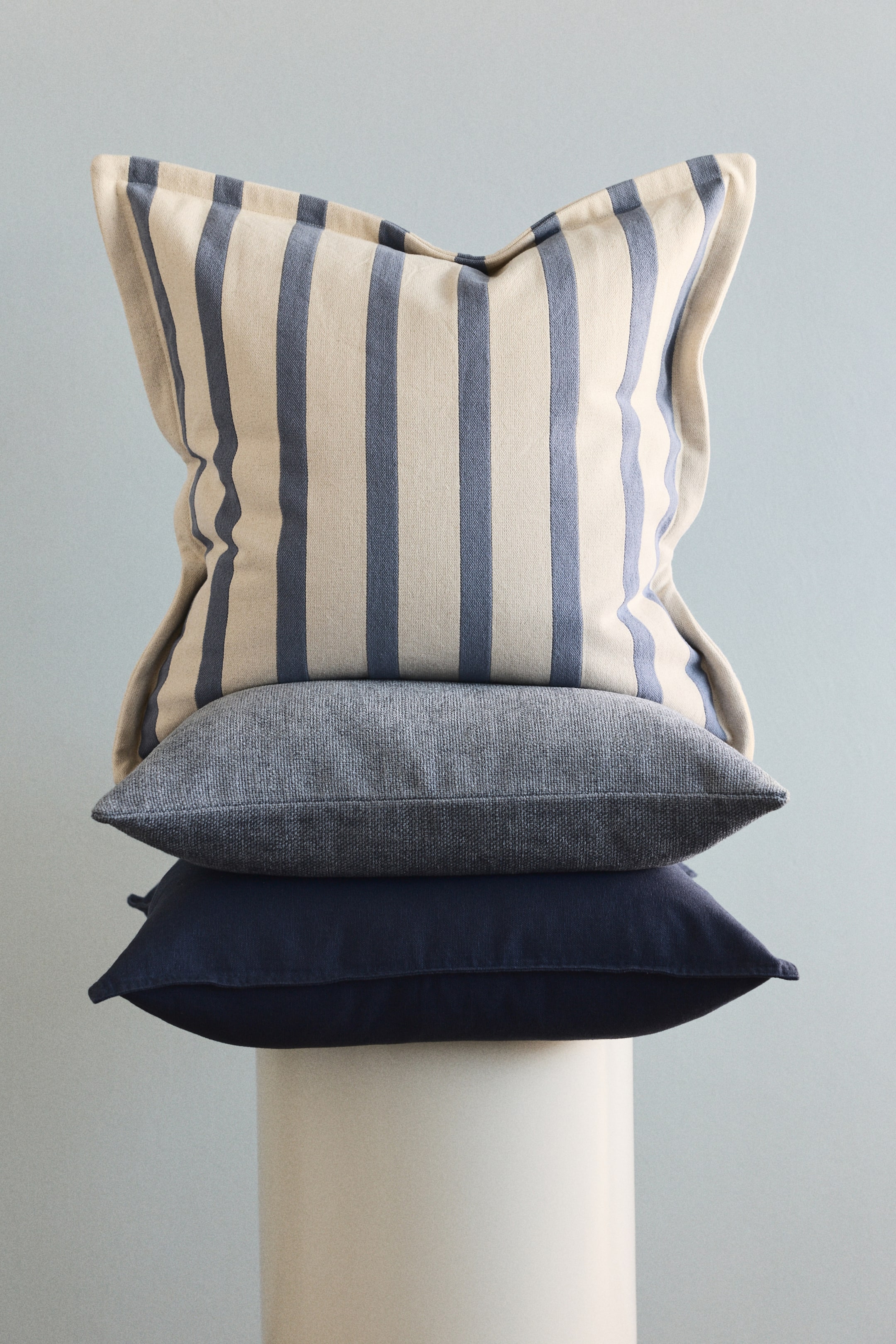 View larger image: Three pillows are stacked, facing forward. The top pillow showcases cream and blue vertical stripes, appearing plush, while the middle pillow features a textured grey-blue fabric, and the bottom pillow displays a smooth dark navy blue.
