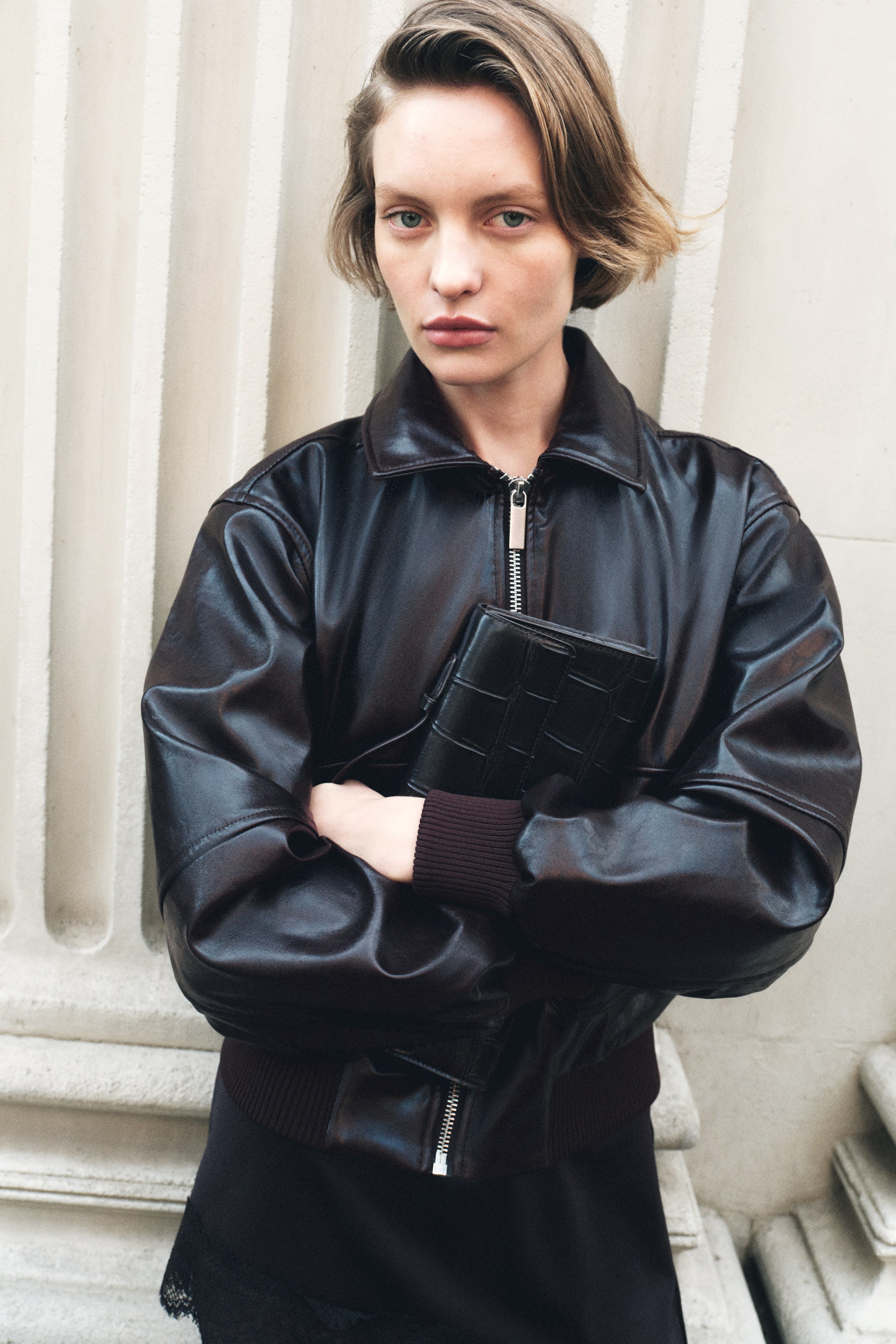View larger image: The model is wearing a short jacket in dark brown coated fabric, showcasing a classic collar, a silver-toned front zip, ribbed cuffs, and a ribbed hem. With arms crossed, the model holds a black textured clutch bag against the jacket.