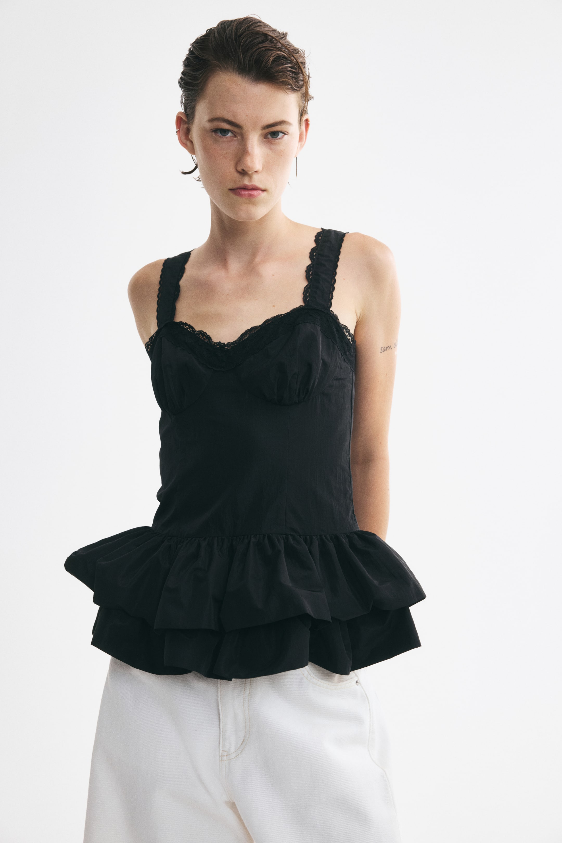 Grotere afbeelding bekijken: Een zwart, strak nylon topje is van voren te zien, met een aansluitend lijfje met gerimpelde, gevormde cups en een sweetheart-halslijn afgewerkt met fijn zwart kant. Het topje heeft brede schouderbandjes met kanten afwerking en loopt vanaf de taille uit in een volumineuze, dubbellaagse volantszoom.