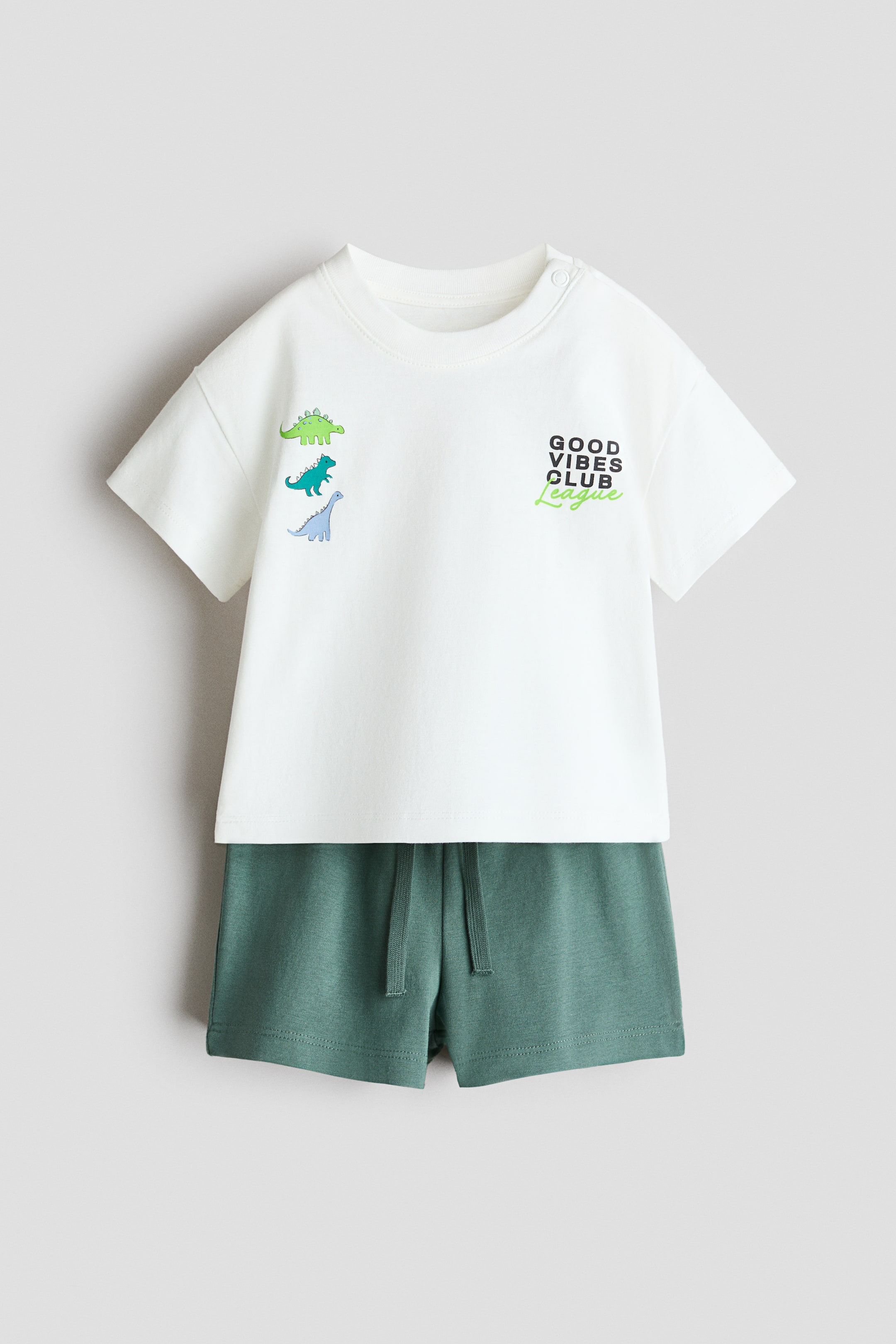 View larger image: Presenting a 2-piece cotton set. The off-white short-sleeved t-shirt features a crew neck with snap buttons, three dinosaur appliqués, and the text "GOOD VIBES CLUB League." Complementing the look, the coordinating dark green shorts come with a drawstring waist.